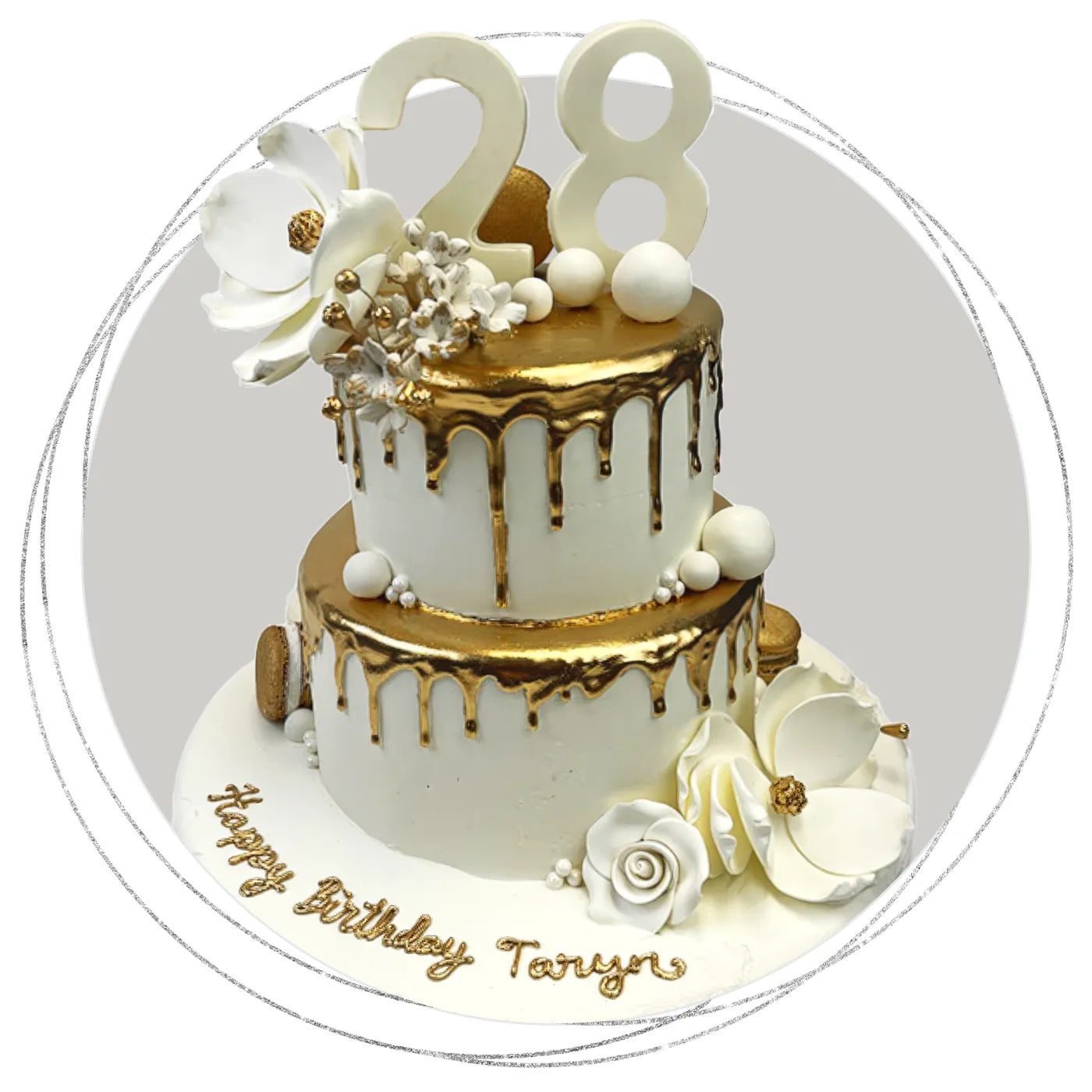 Two-tier white and gold drip birthday cake with sugar flowers, macarons, and “28” topper.