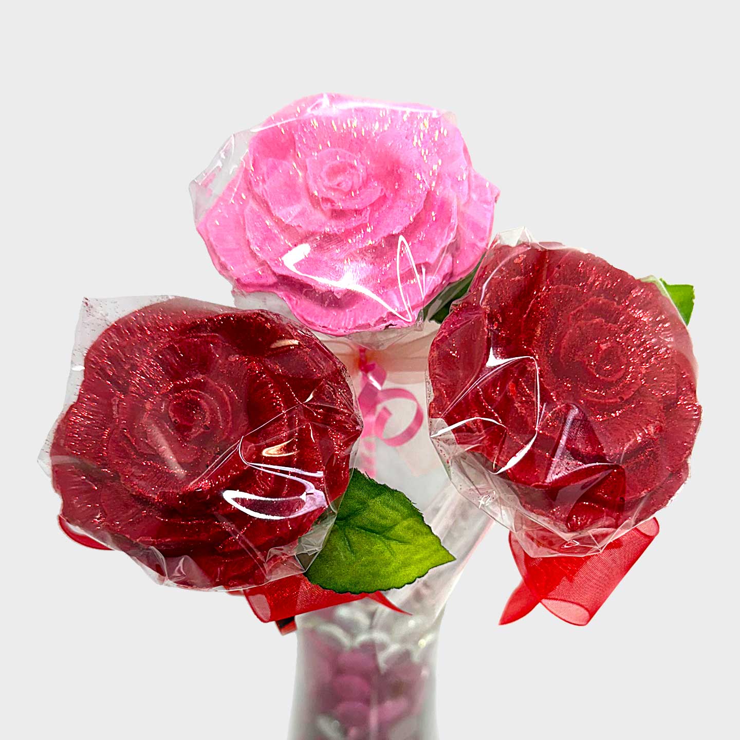 A pink and two red chocolate roses with green faux leaves, each elegantly wrapped in clear cellophane and tied with satin ribbon