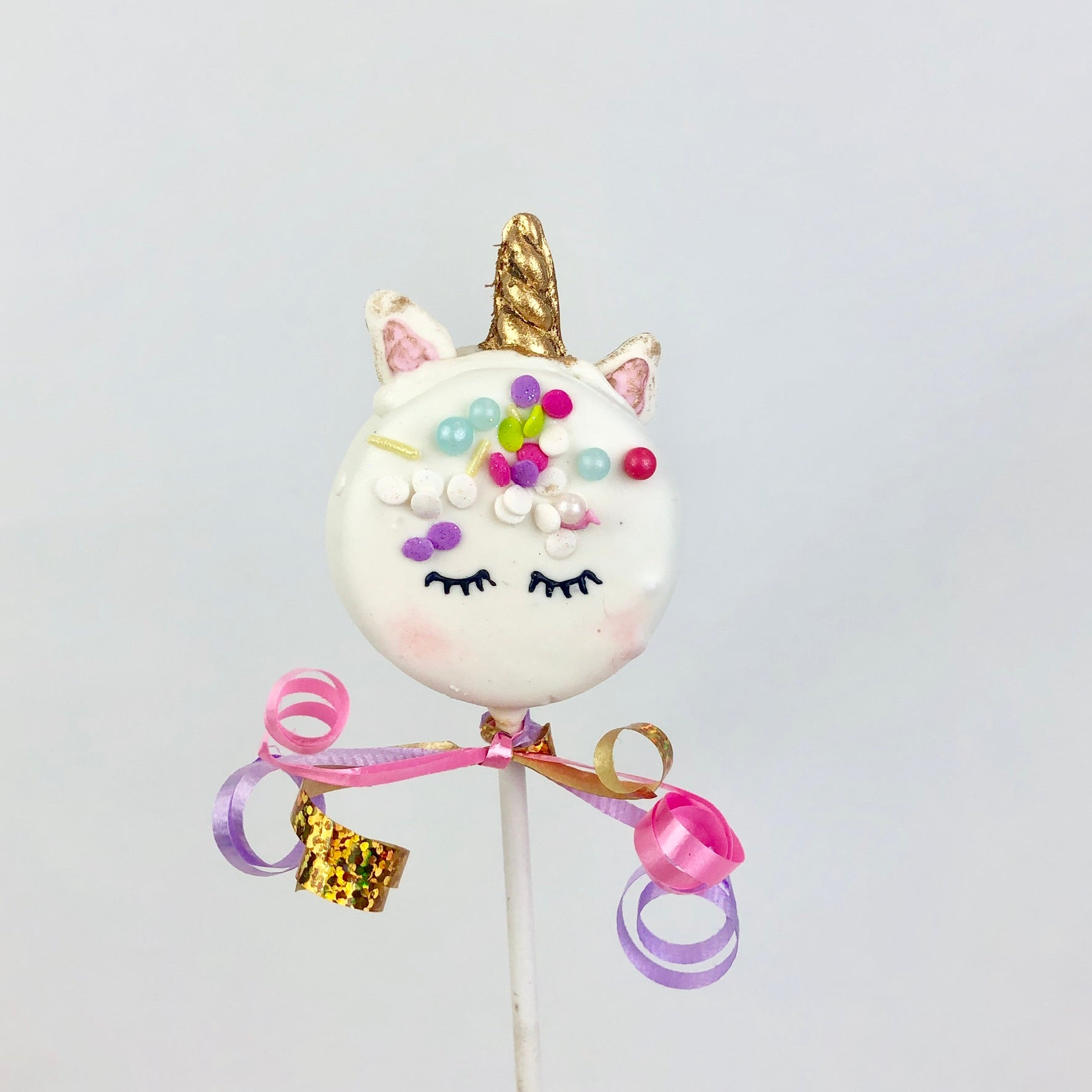 Chocolate covered Oreo decorated like a unicorn. The stick is wrapped with ribbons.