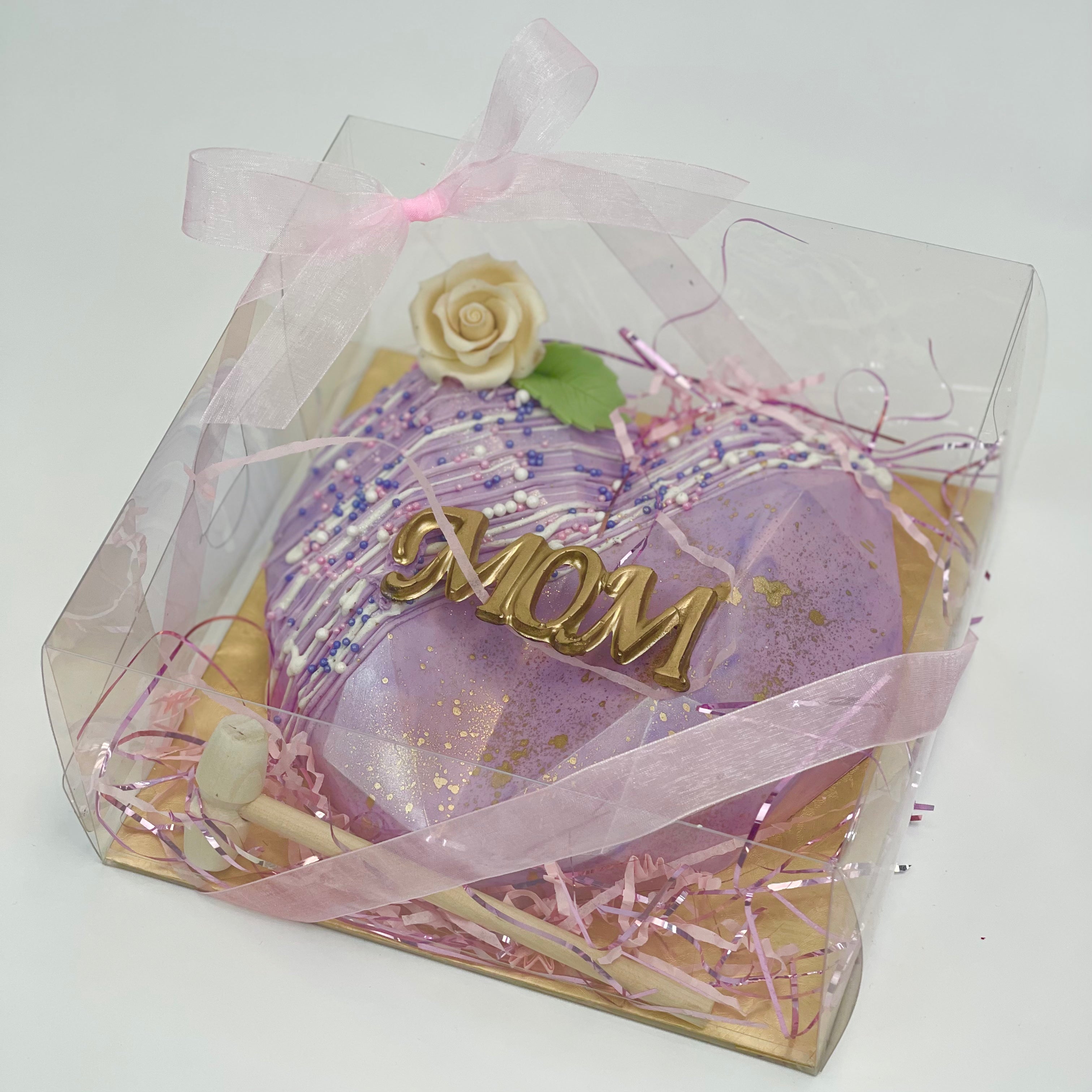 A purple, candy-filled chocolate heart smash piñata rests on a golden base, and a wooden mallet, inside a clear box, wrapped with a pink ribbon. The top is decorated with 'Mom' written in gold, a rose, and colorful sprinkles.