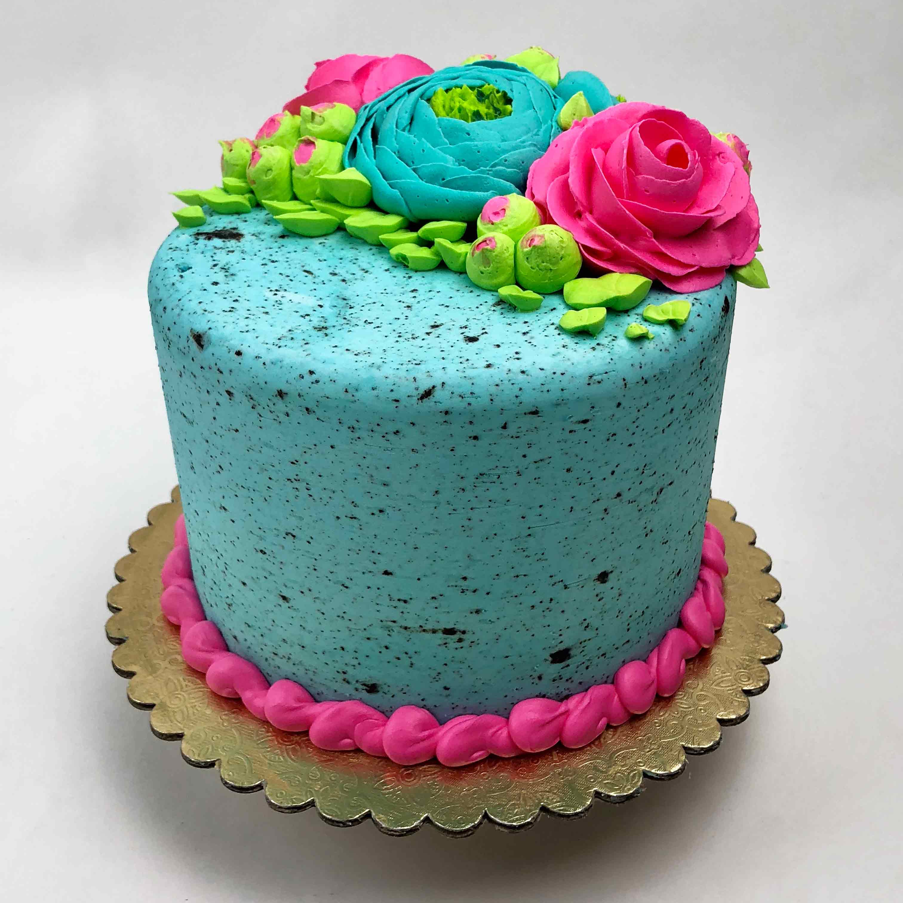 A blue Easter buttercream cake, decorated with pink, blue and green flowers on top. The cake has pink piping around the base and sits on a golden base.