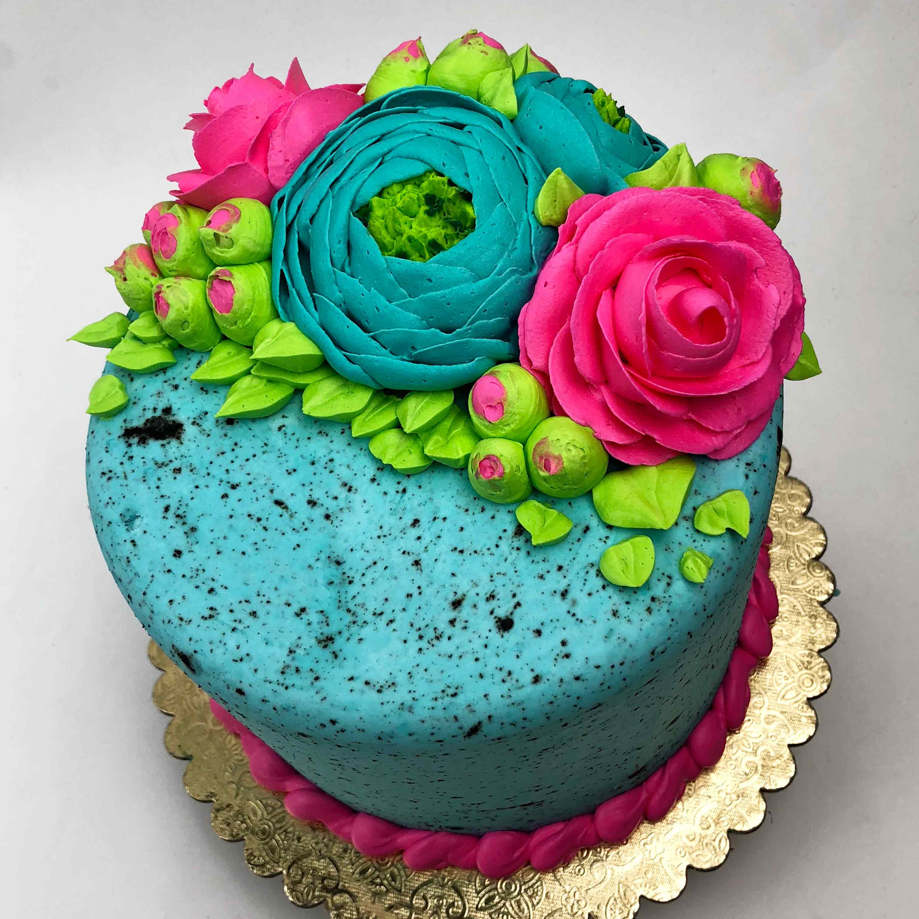The top view of a blue Easter buttercream cake, decorated with pink, blue and green flowers on top. The cake has pink piping around the base and sits on a golden base.