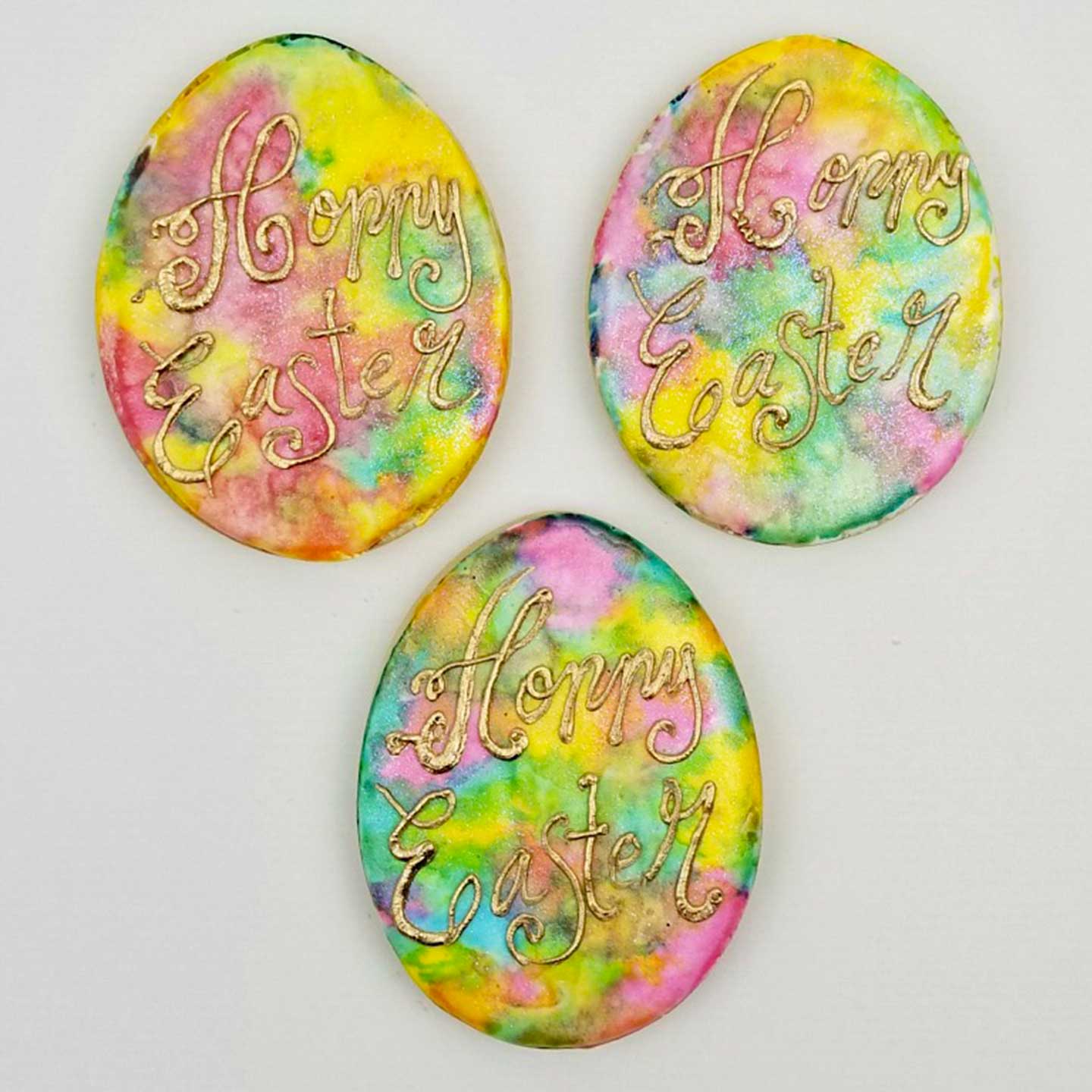 Three sugar cookies decorated with pastel watercolor designs, each featuring a golden color "Hoppy Easter" message.