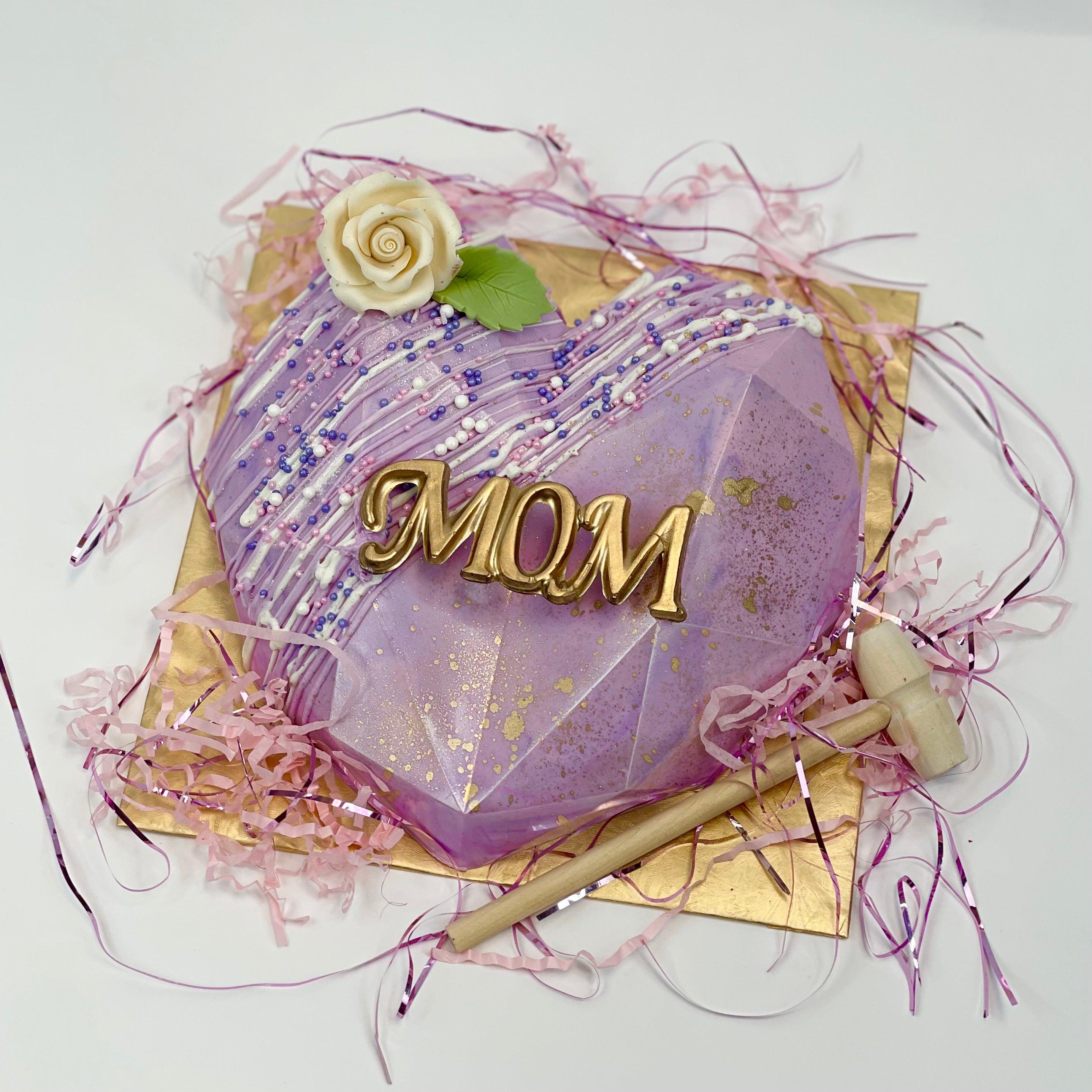 A purple, candy-filled chocolate heart smash piñata rests on a golden base, and a wooden mallet. The top is decorated with 'Mom' written in gold, a rose, and colorful sprinkles.