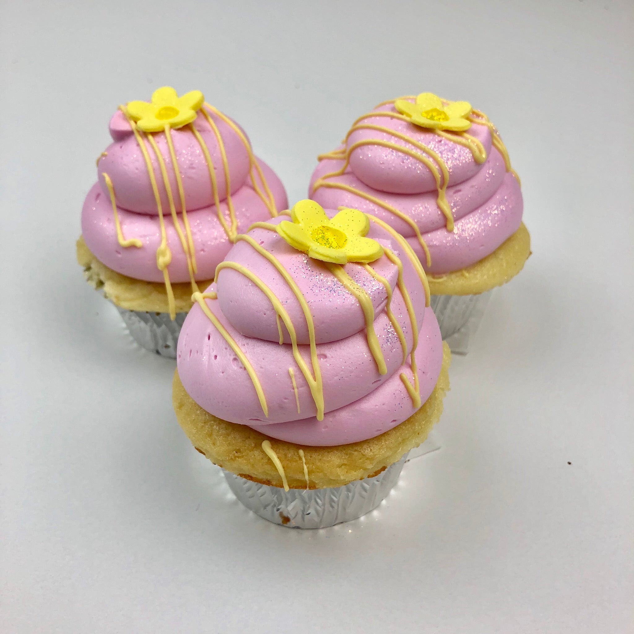 Three cupcakes with pink frosting, a yellow fondant flower on top, drizzled with lemon chocolate, and edible glitter