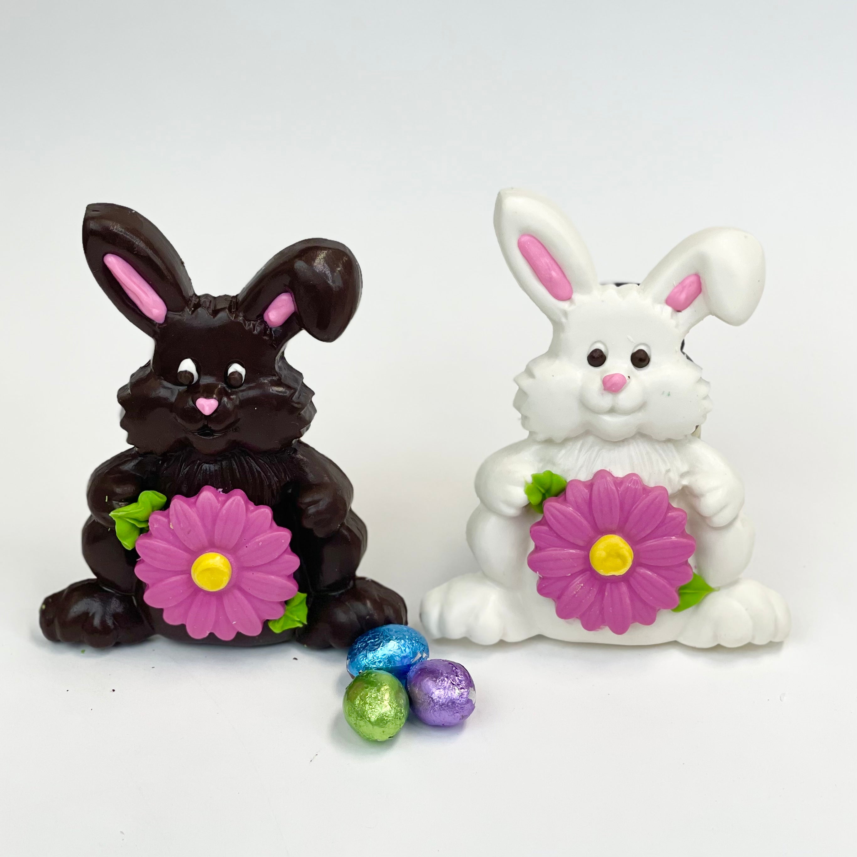 A dark brown and white chocolate bunny sitting next to each other, each holding a pink flower with a yellow center and green leaves. In front of them are three mini Easter egg chocolates wrapped in blue, green, and purple metallic paper.