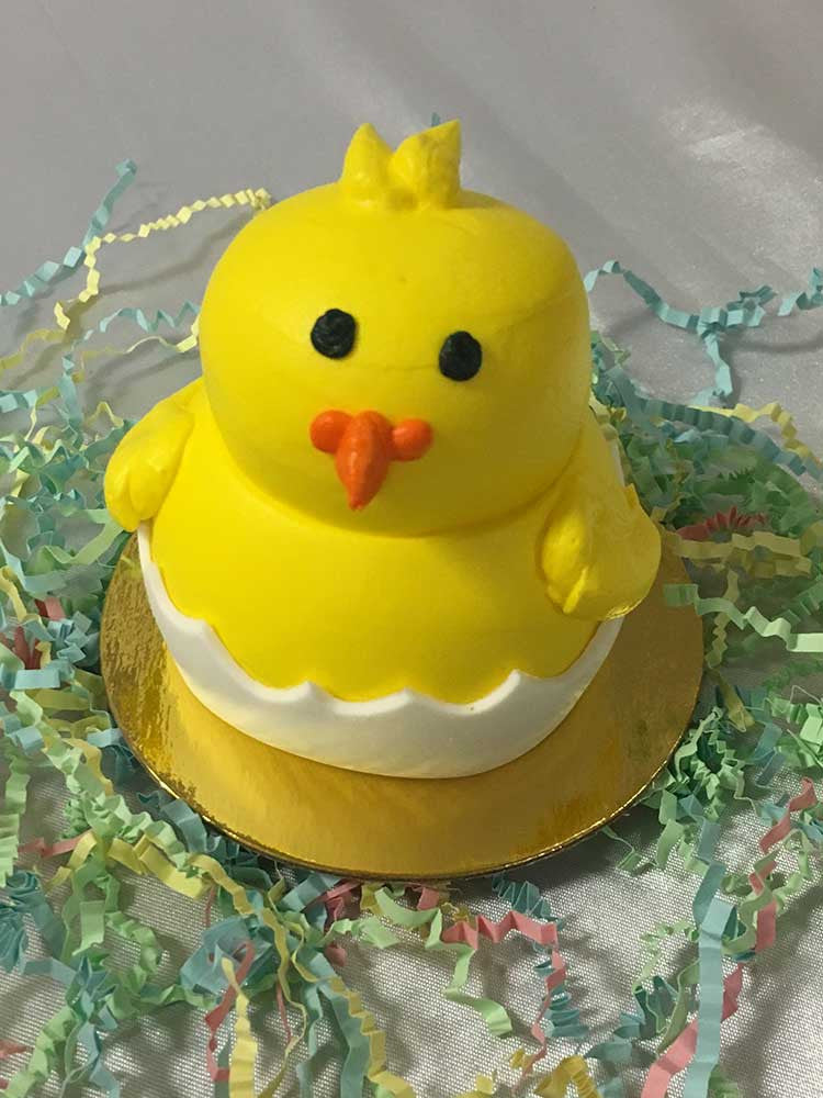 A yellow chick inside half of an egg shell Easter 3D Cupcake sitting on top of a golden base. There are some pastel colored crinkled paper around the cupcake.