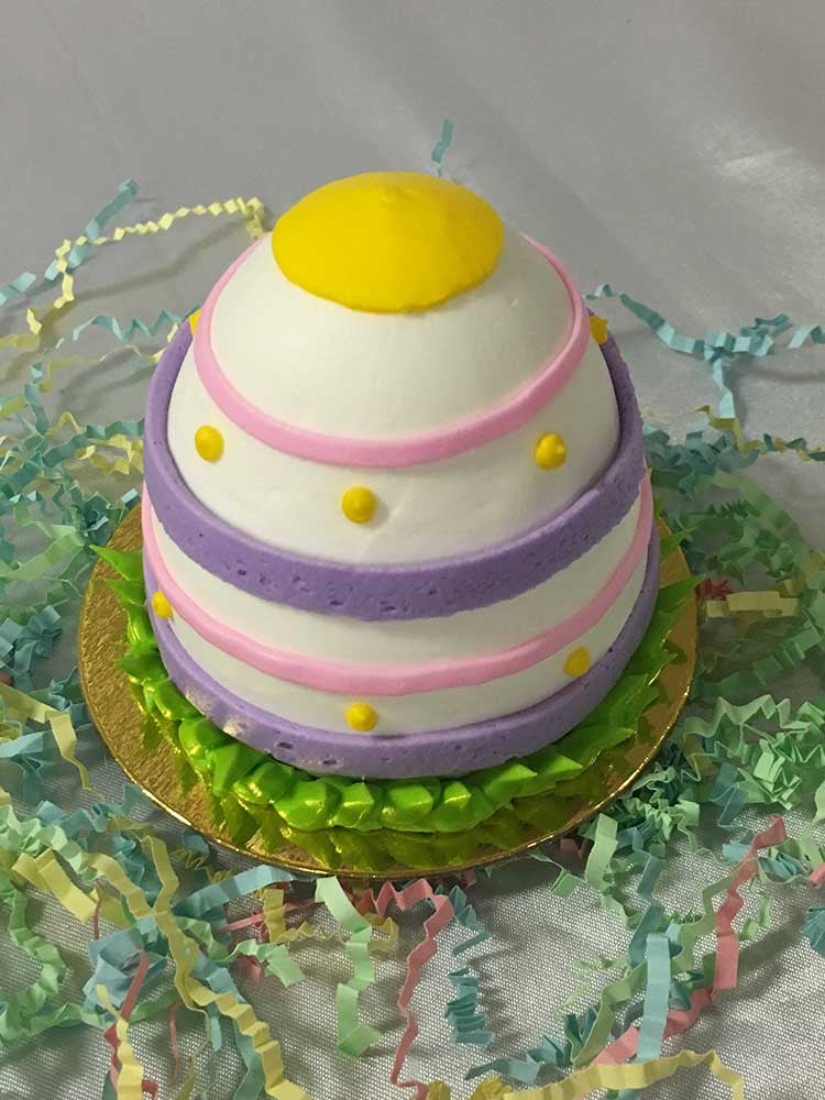 A white Easter Egg 3D Cupcake sitting on top of a golden base. The egg is decorated with yellow, pink, purple, and green piping around it. There are some pastel colored crinkled paper around the cupcake.