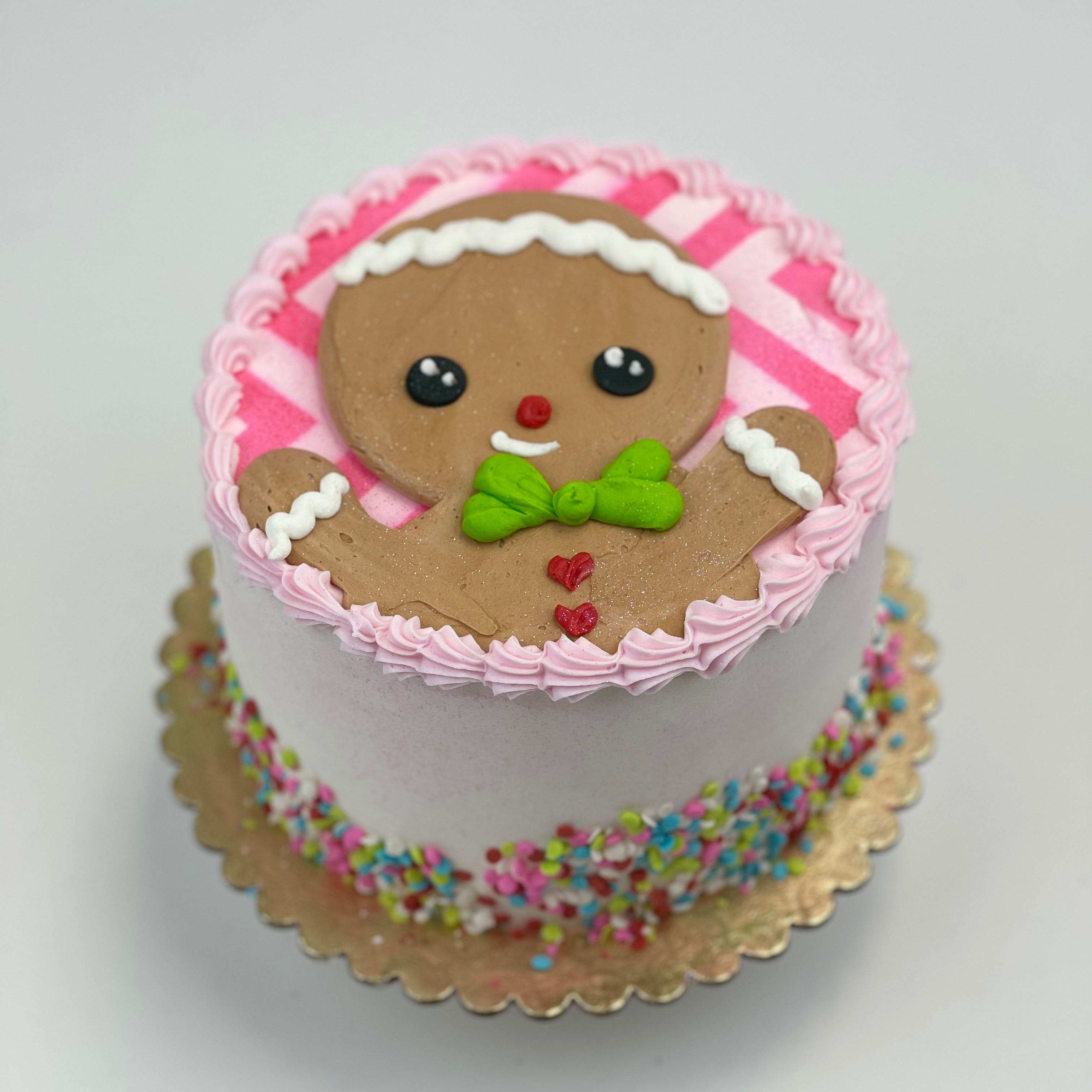 The Gingerbread Christmas Cake is iced with pink buttercream. The gingerbread man is wearing a green bow, has a red note, and red heart-shaped buttons.