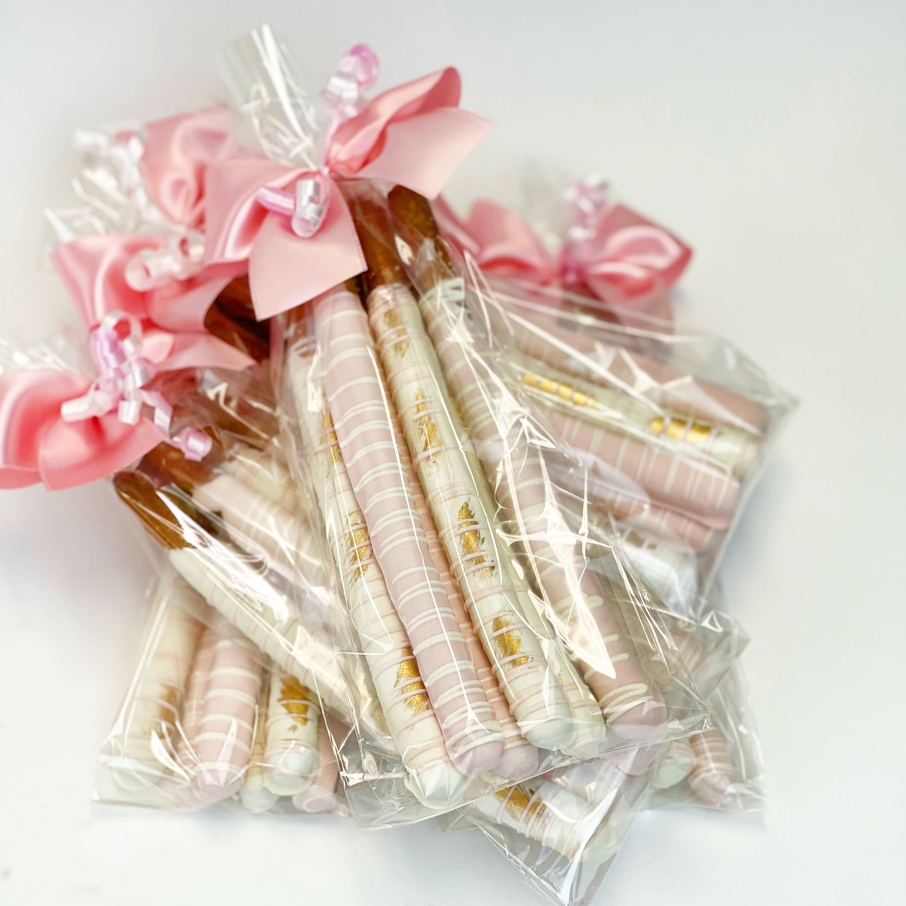 Clear gift bags filled with six chocolate-covered pretzels, three dipped in light pink with white drizzle, and three dipped in white with pink and gold drizzle, tied with pink bows.