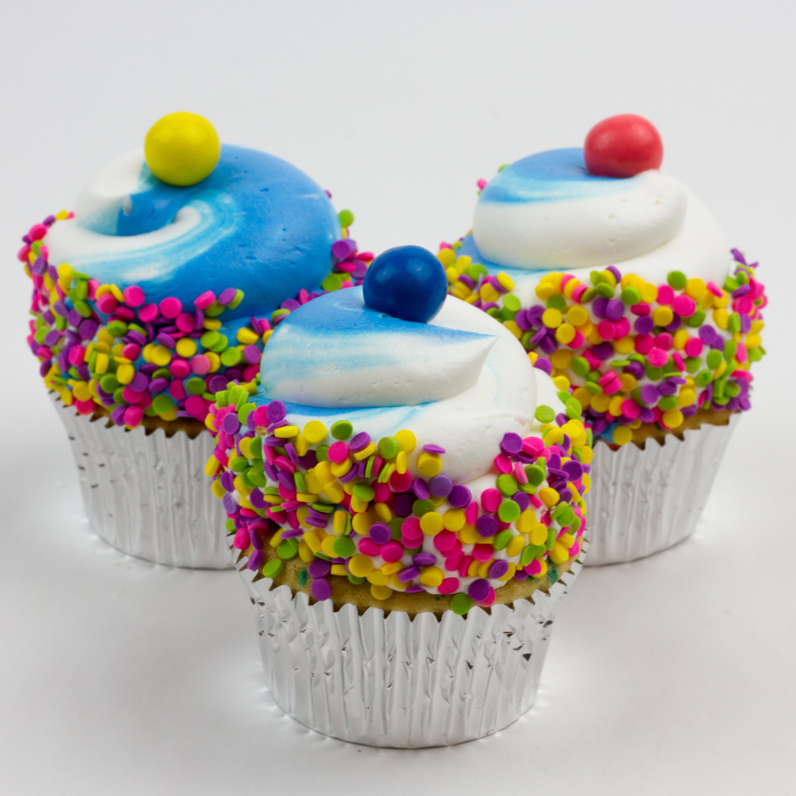 Three Blue Funfetti Cupcakes topped with a blue and white buttercream swirl and each are garnished with sprinkles and yellow, blue, pink bubblegum balls