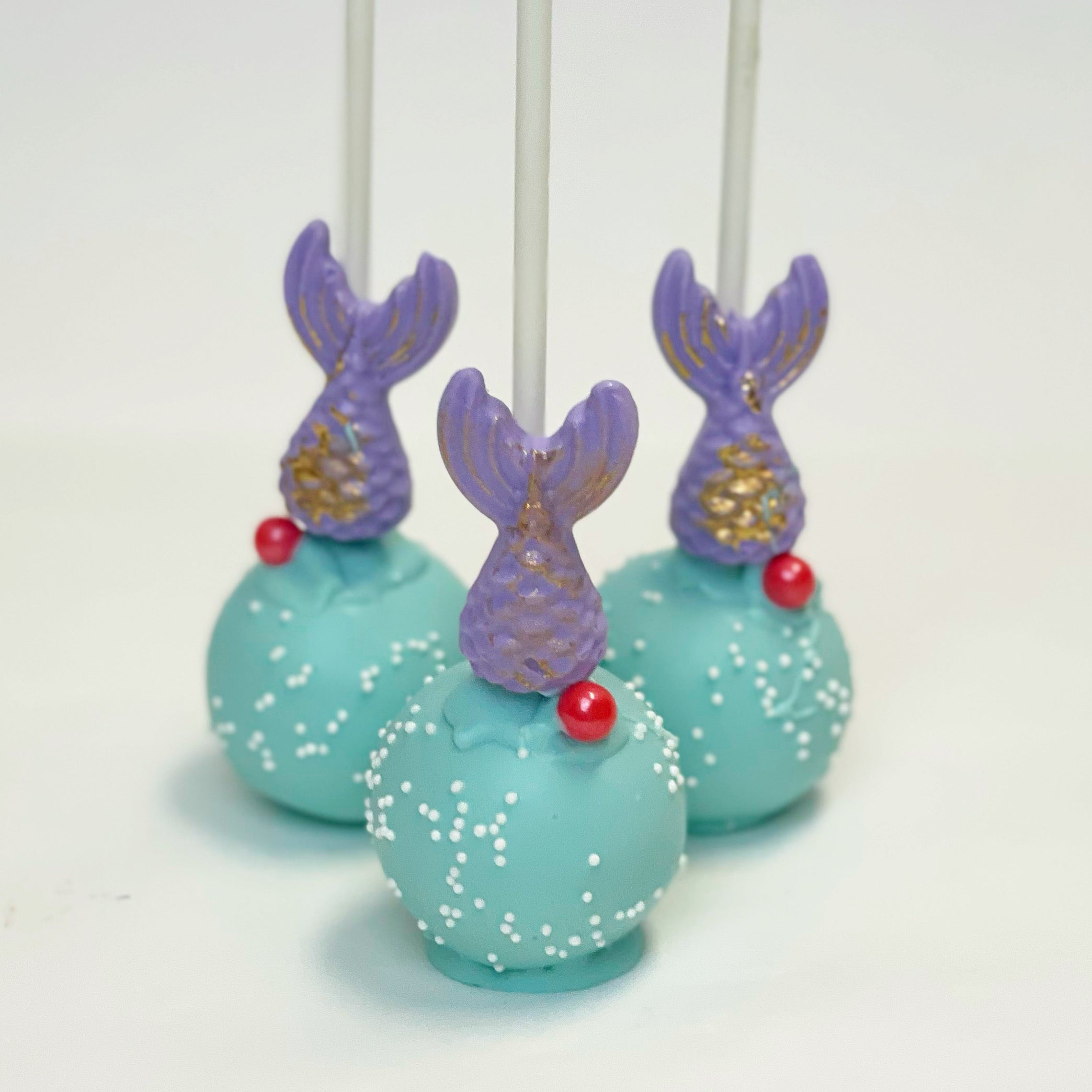 Three mermaid cake pops with aqua bodies and small white sprinkles, purple tails with gold accents, and a small red candy detail