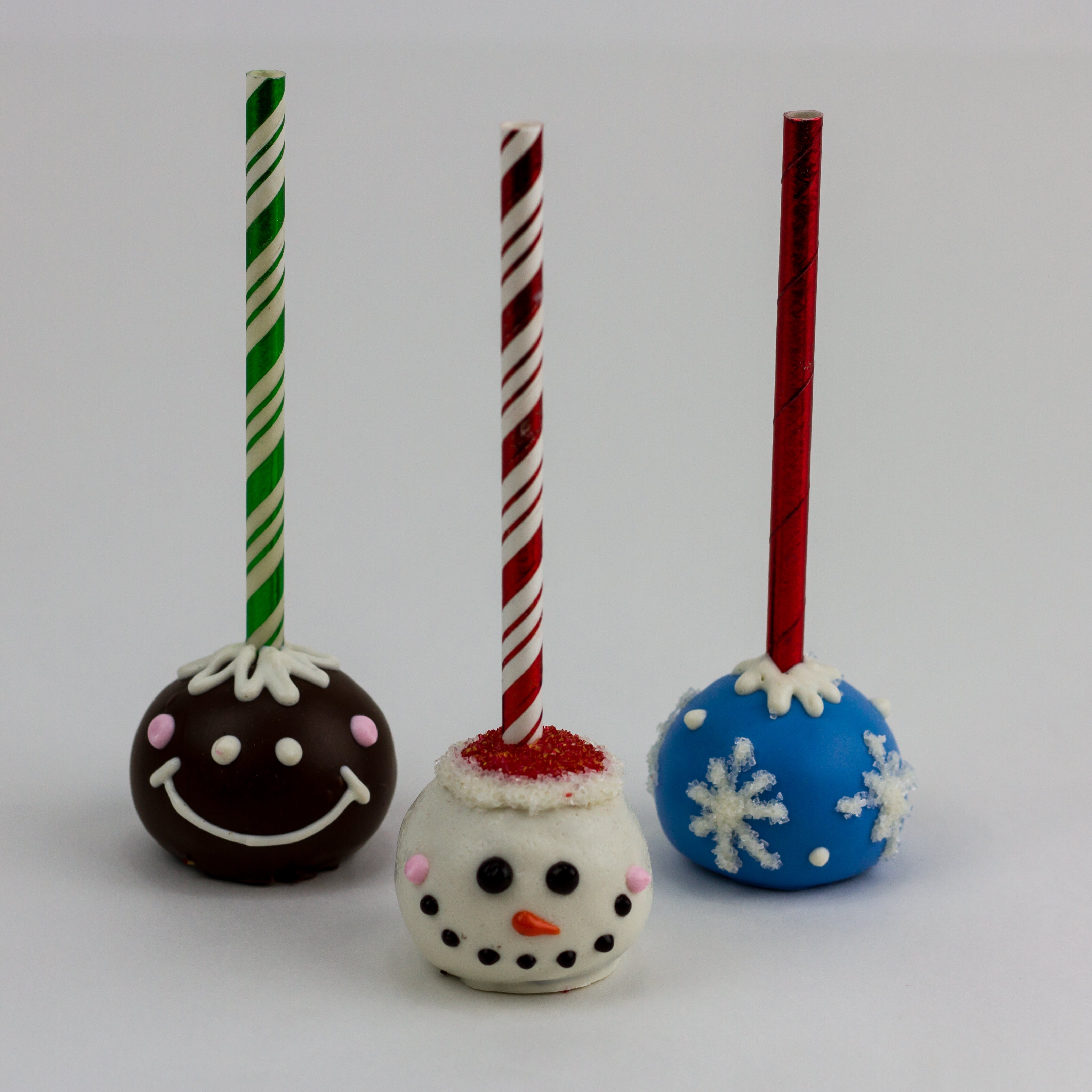 A smiling brown gingerbread character, a smiling snowman with a red hat, and a blue and white snowflake cake pop.