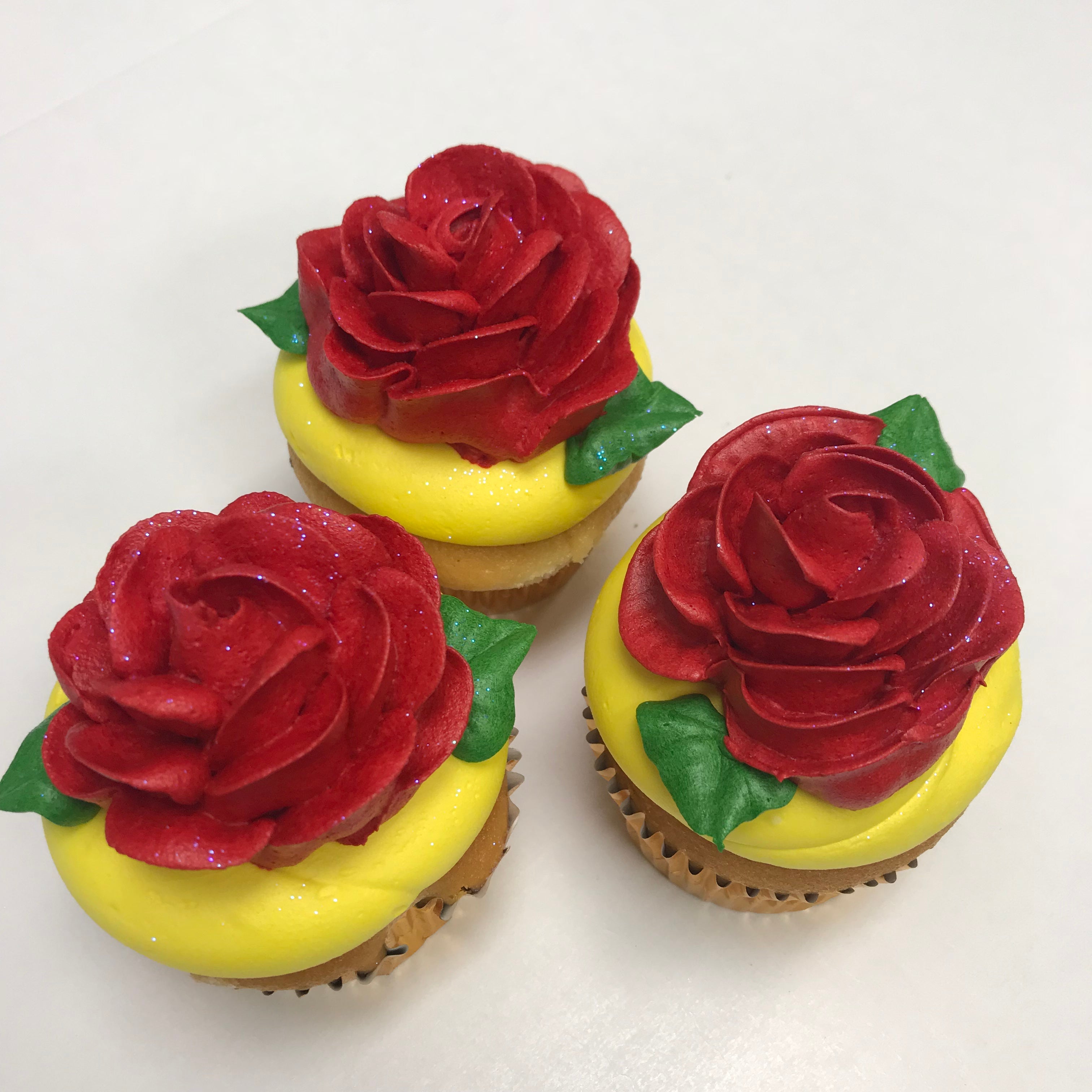 Three cupcakes with yellow frosting and a large red buttercream rose on top, garnished with green leaves.