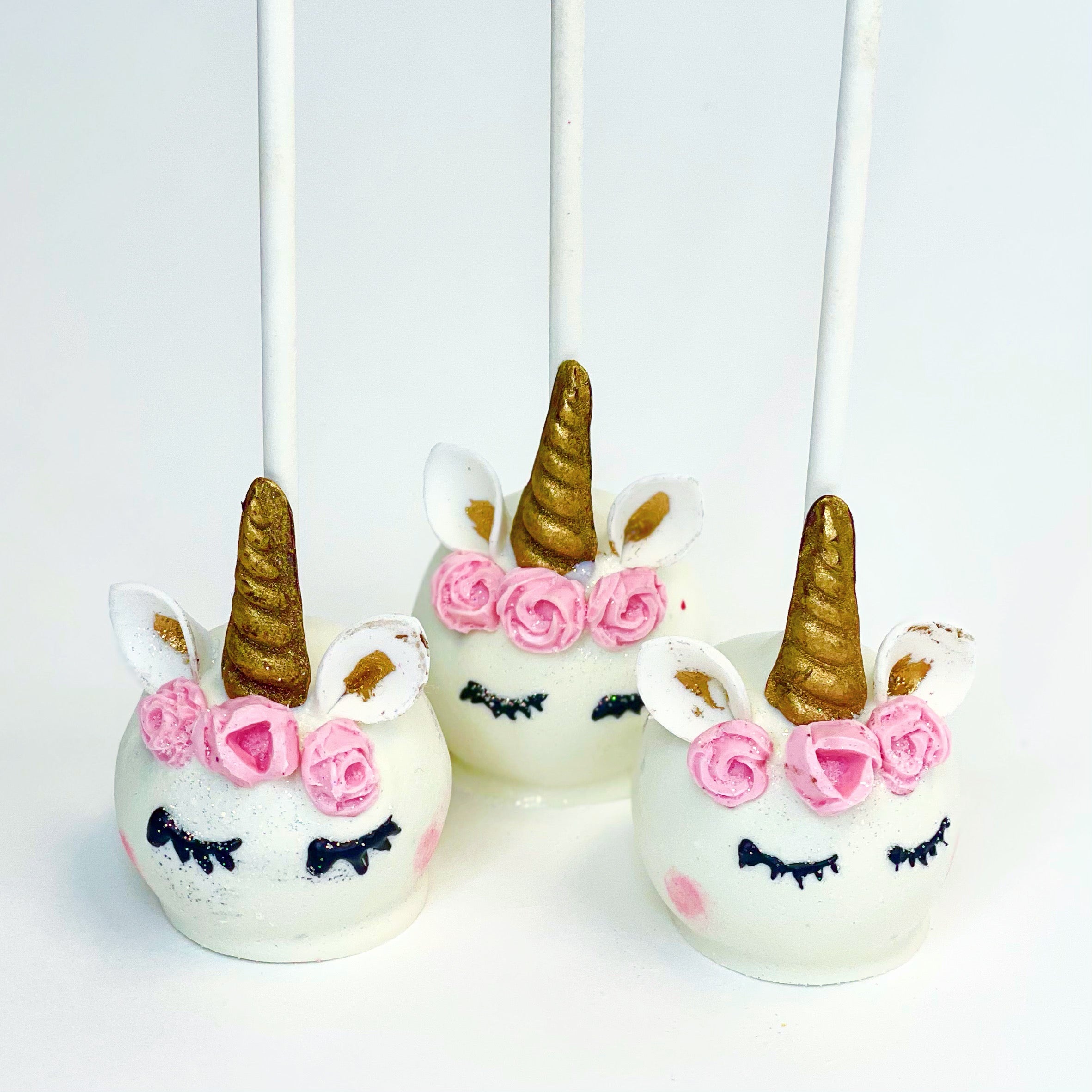 Three white unicorn cake pops, each with a golden horn, golden inner ears, three pink flowers on their heads, black closed eyes, and pink cheeks