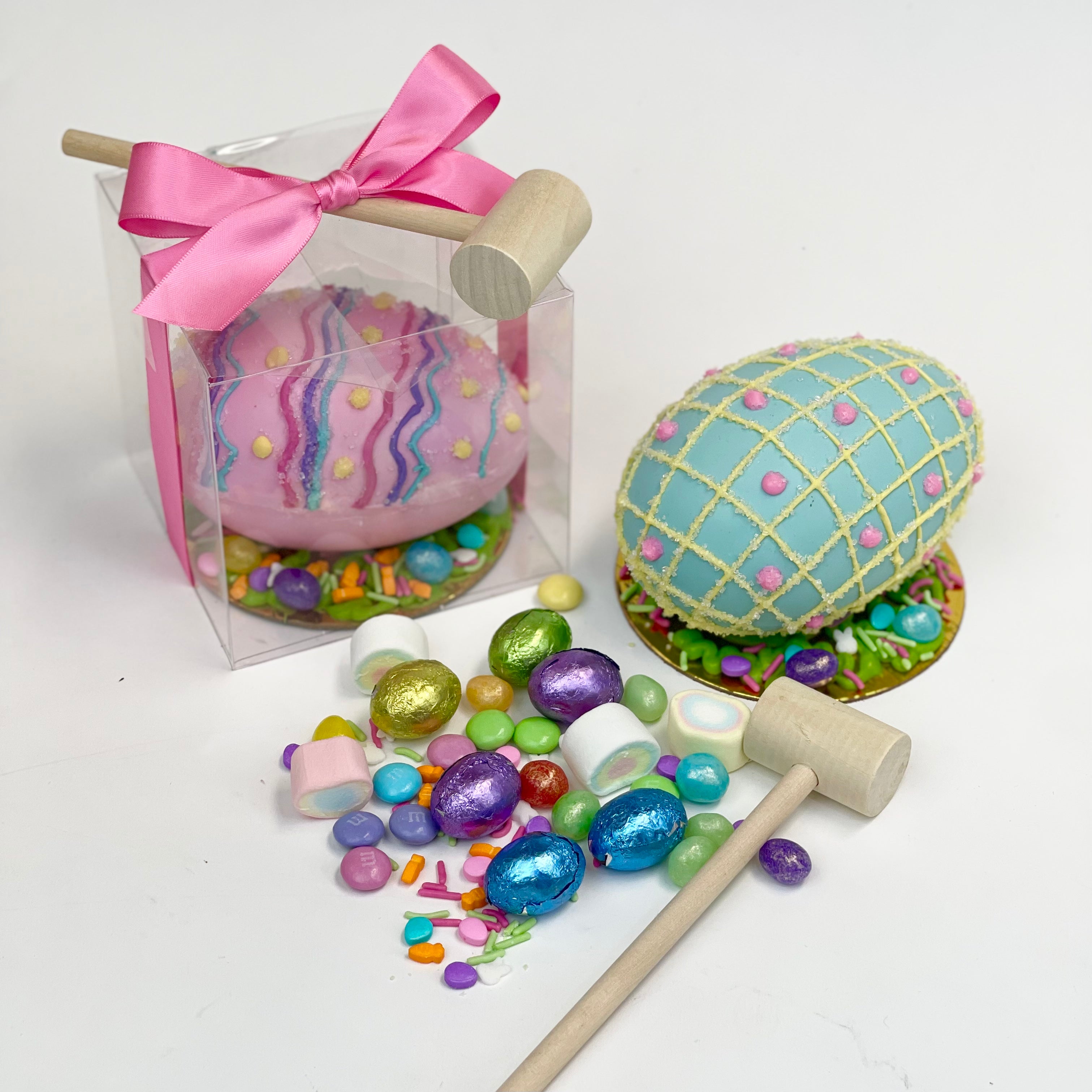 A decorated pink piñata Easter egg inside a clear box, wrapped with a pink ribbon and accompanied by a mallet. Next to it, a decorated blue piñata Easter egg sits on a gold base, surrounded by Easter candy and a mallet.