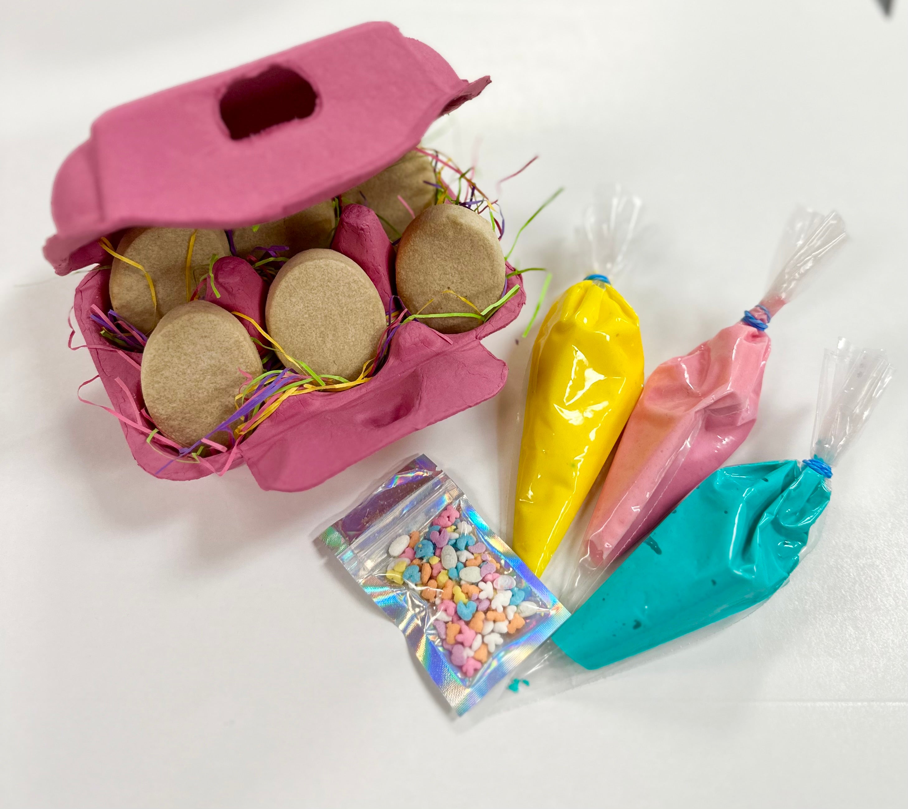 6 Easter egg-shaped sugar cookies inside a pink colored egg cartoon next to 3 small bags of yellow, pink, and blue royal icing and a bag of sprinkles.