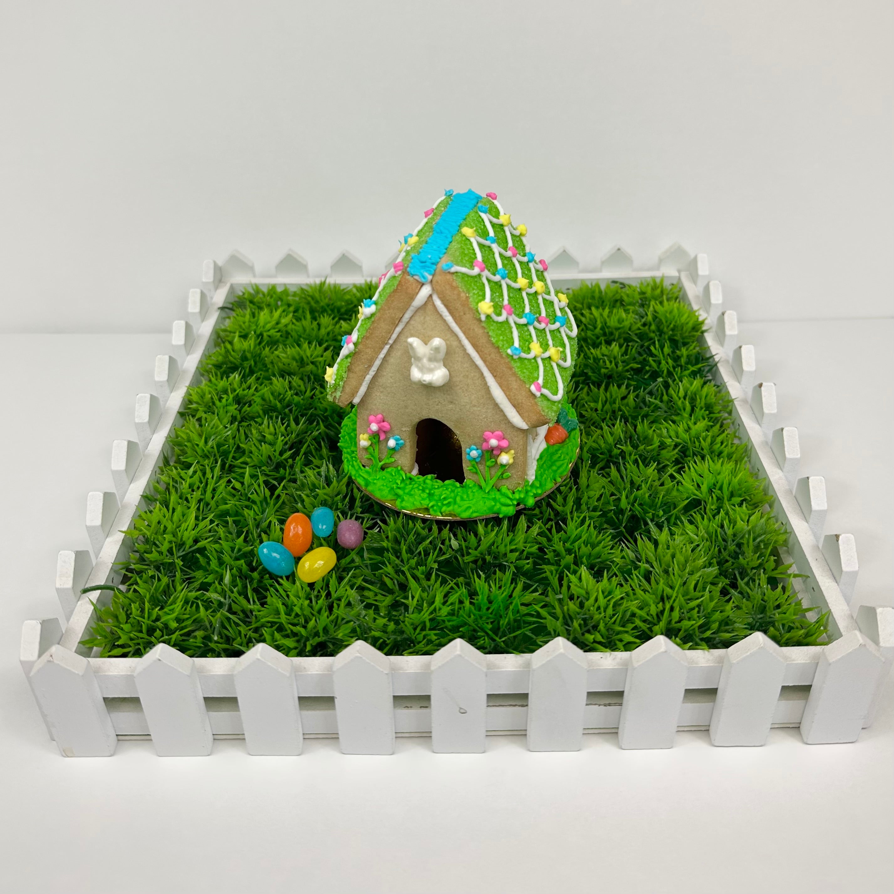 A mini Easter house make of sugar cookies, nestled inside a box filled with decorative plastic grass, some jelly beans, and a white picket fence.