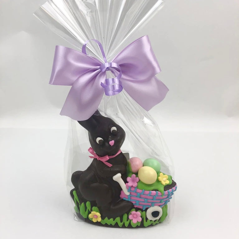 A solid chocolate Easter bunny standing next to an Easter basket with flowers and three colorful mini easter eggs, all wrapped in cello with a purple satin bow.