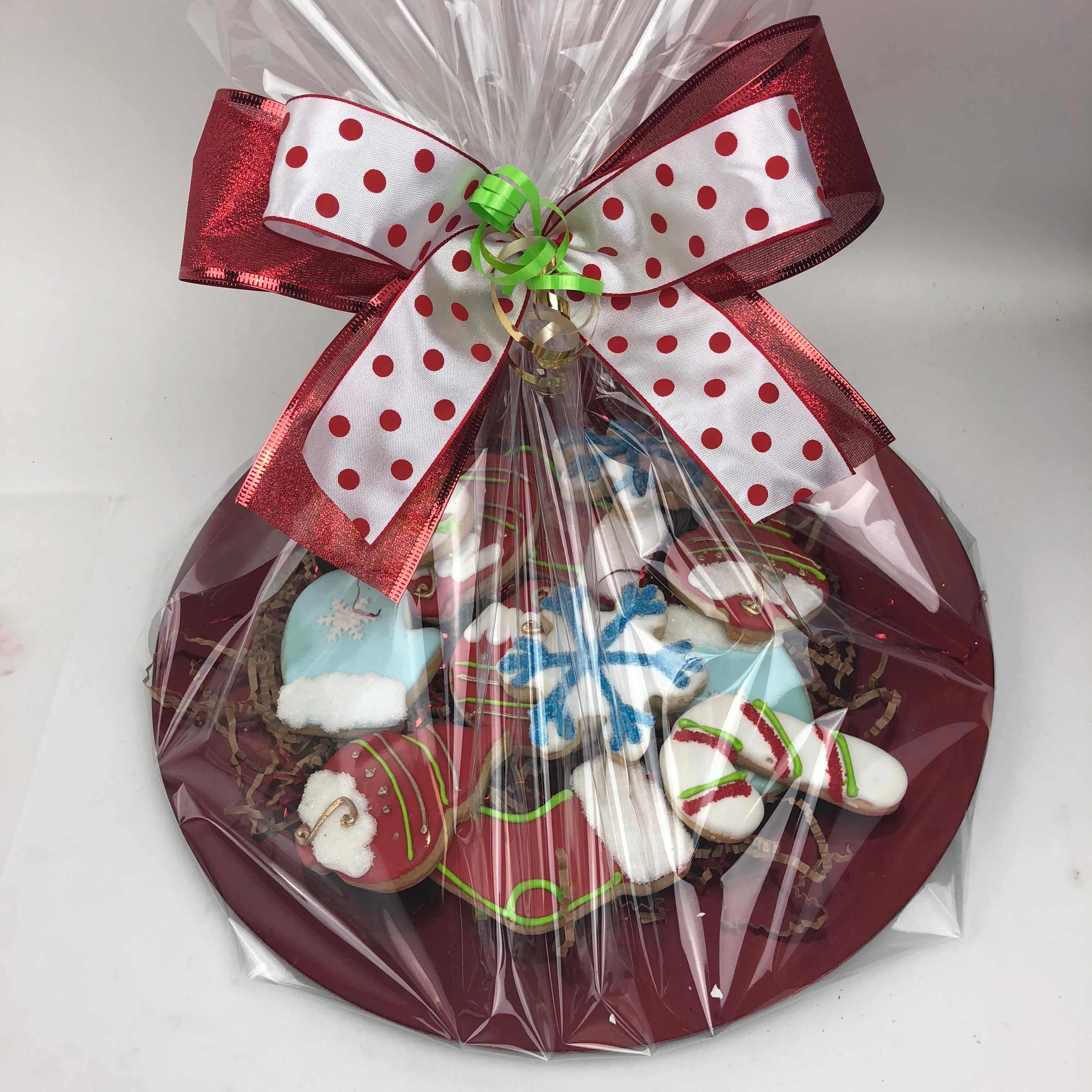 A platter of 12 assorted medium decorated sugar cookies in the shape of ornament, mittens, stocking, candy cane, and snowflake. They are all wrapped up in cello with a pretty red, white, and green satin bow.
