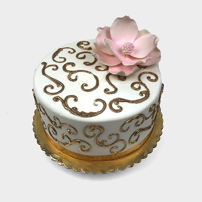 White cake with gold scrolls and a pink magnolia flower on top.