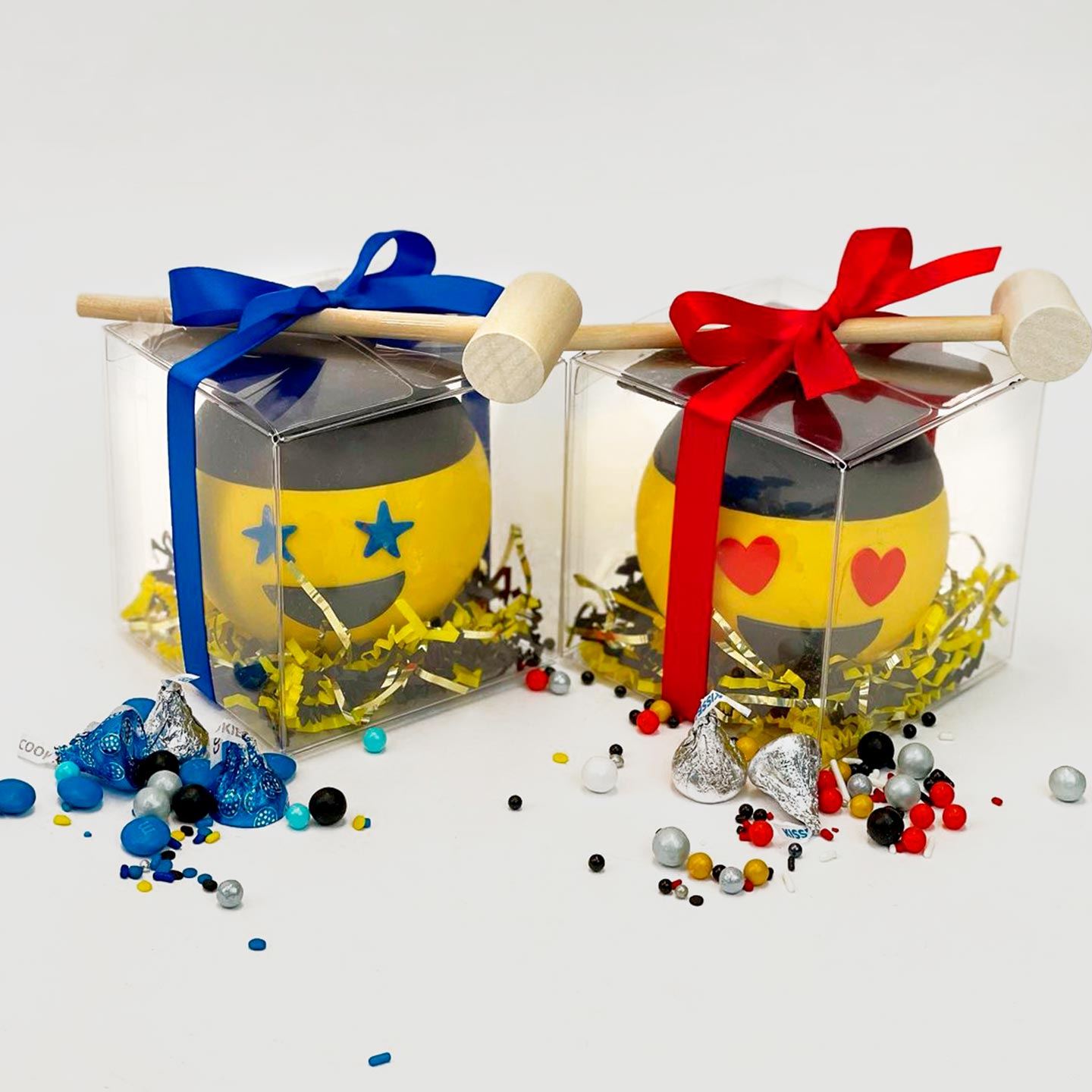 Two chocolate happy faces wearing a graduation cap. Each is in a clear box. The box is wrapped with a bow and a mallet. Assorted candies are spread on the outside to show the content. There are two options: blue and red.
