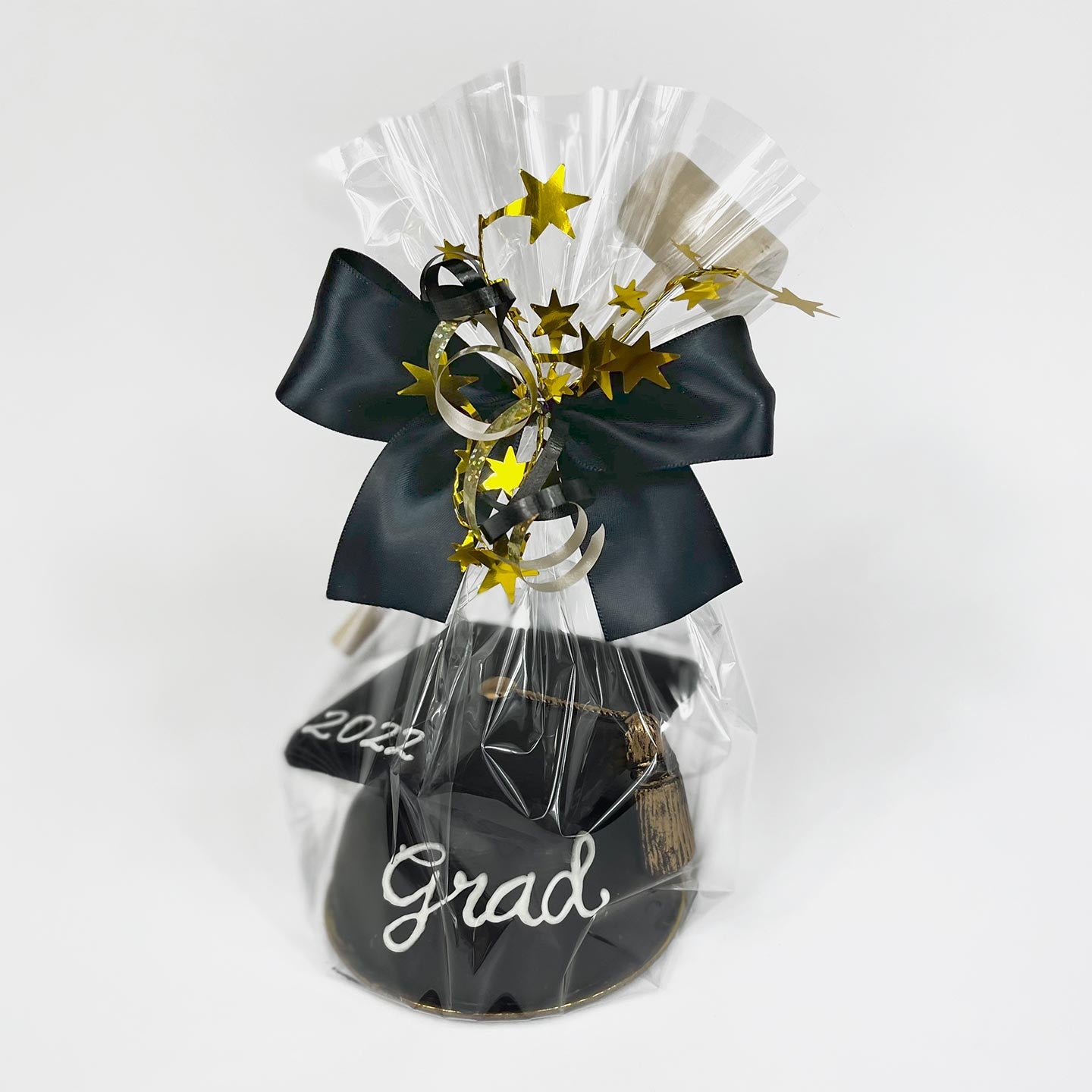 A black chocolate graduation cap is shown wrapped in cellophane with a black bow, silver ribbon, and metallic gold stars.