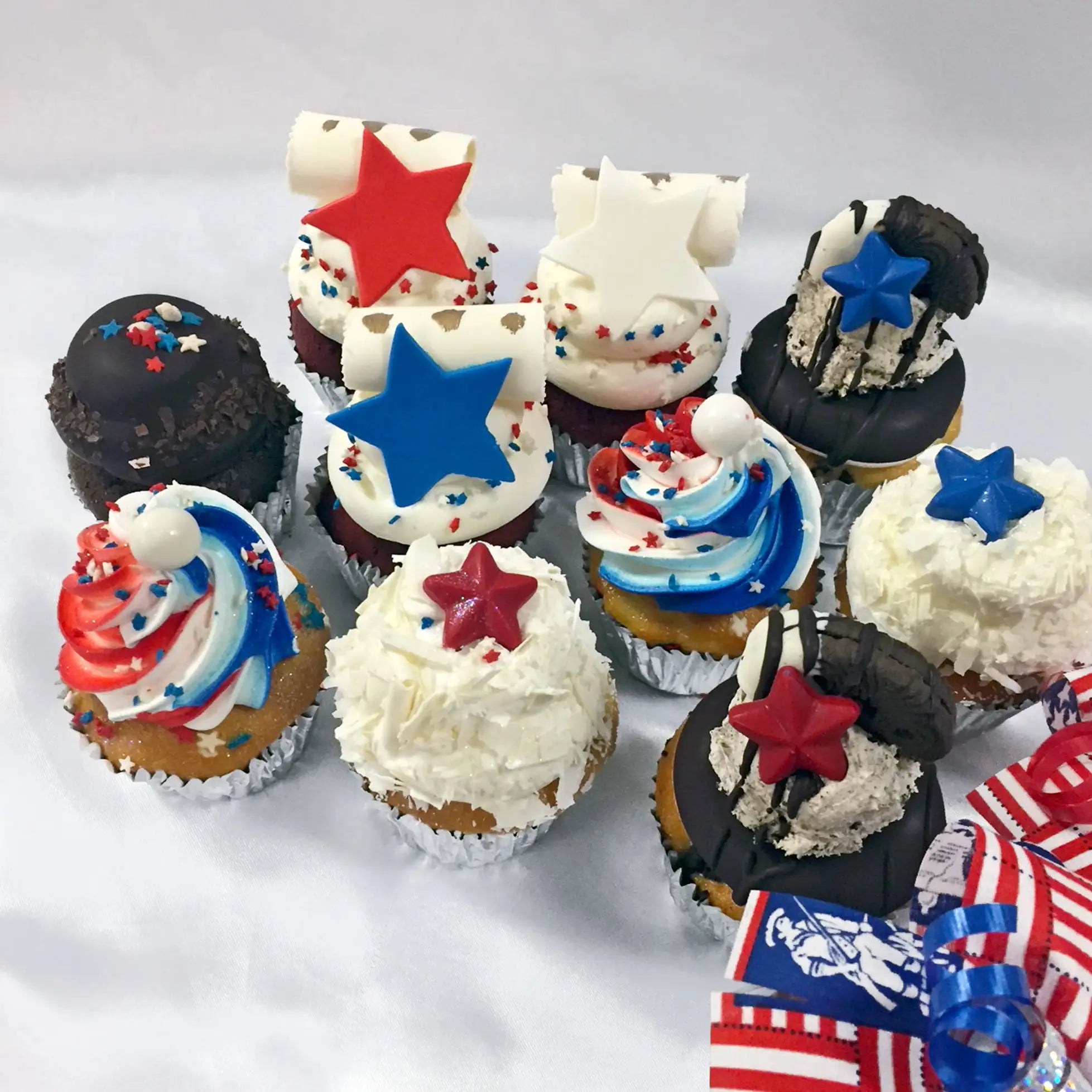 Nine 4th of July–themed assorted cupcakes arranged on a light surface, each topped with red, white, and blue frosting, sprinkles, star-shaped toppers or scroll decorations.