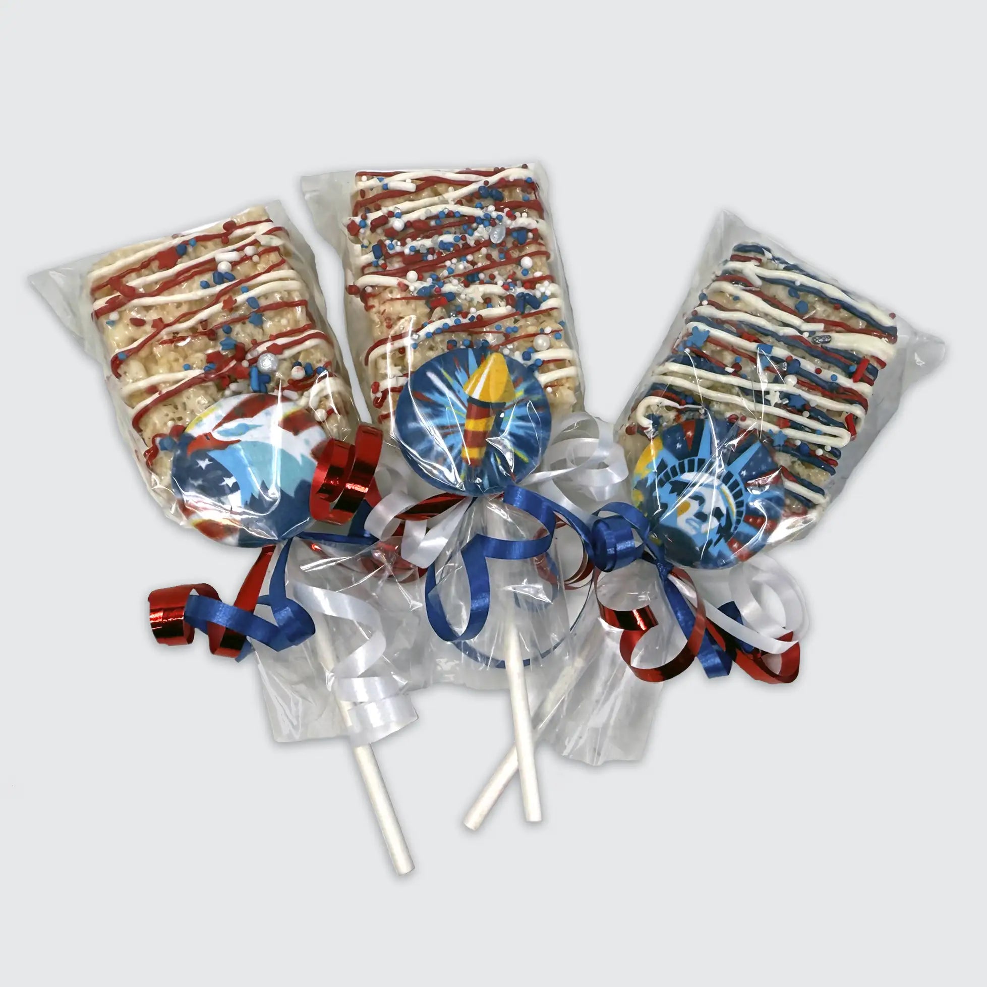 Three Rice Krispy treat pops decorated with red, white, and blue drizzle and patriotic sprinkles, each wrapped in cello with red, white, and blue curling ribbons and topped with themed chocolate medallions; American Eagle, Firecracker, and Statue of Liberty.