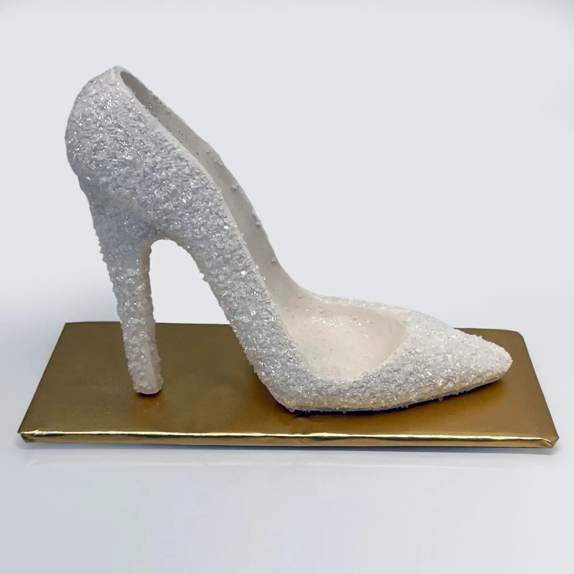 A white chocolate high heel shoe, encrusted with shimmering sugar crystals, set on a chic golden base