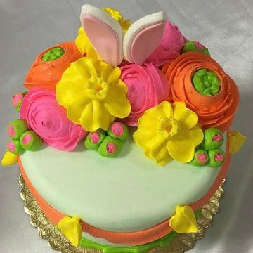 The top view of a pastel green Bunny Ears Floral Cake featuring yellow, pink, orange, and green flowers elegantly arranged on top, with delicate yellow, orange, and green piping embellishing the sides. The cake is displayed on a golden base.