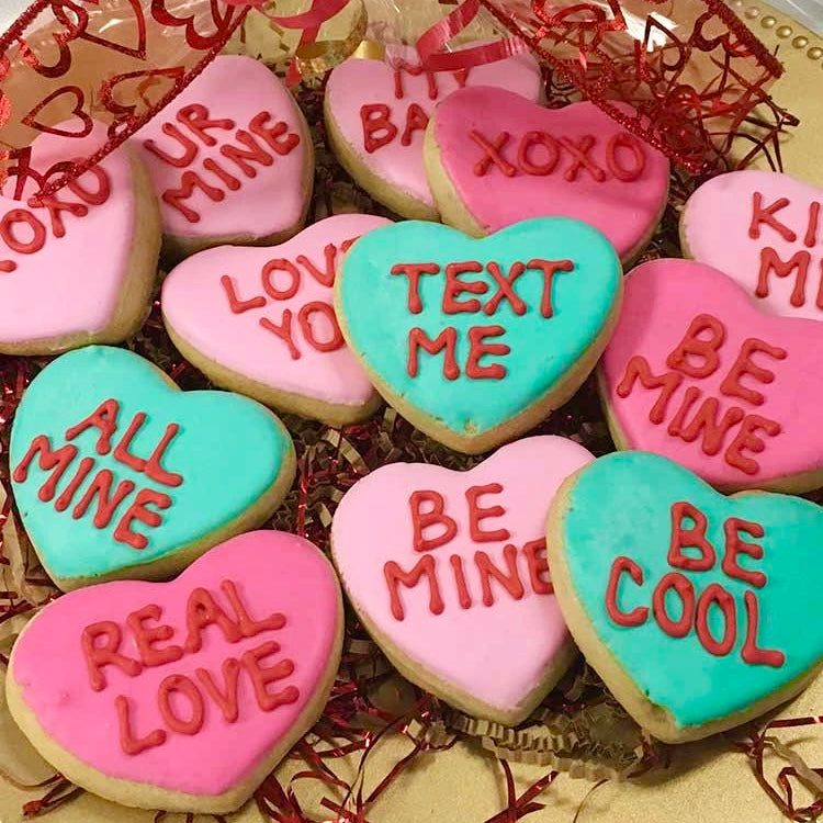 A dozen of ink and teal color heart-shaped sugar cookies decorated with fun Valentine’s Day sayings like "XOXO", "Be Mine", "Be Cool", "All Mine" "Real Love", "Kiss Me", "Love You", "UR Mine" and "Text Me" on them and wrapped on a platter in clear cellophane and tied with a beautiful red bow.