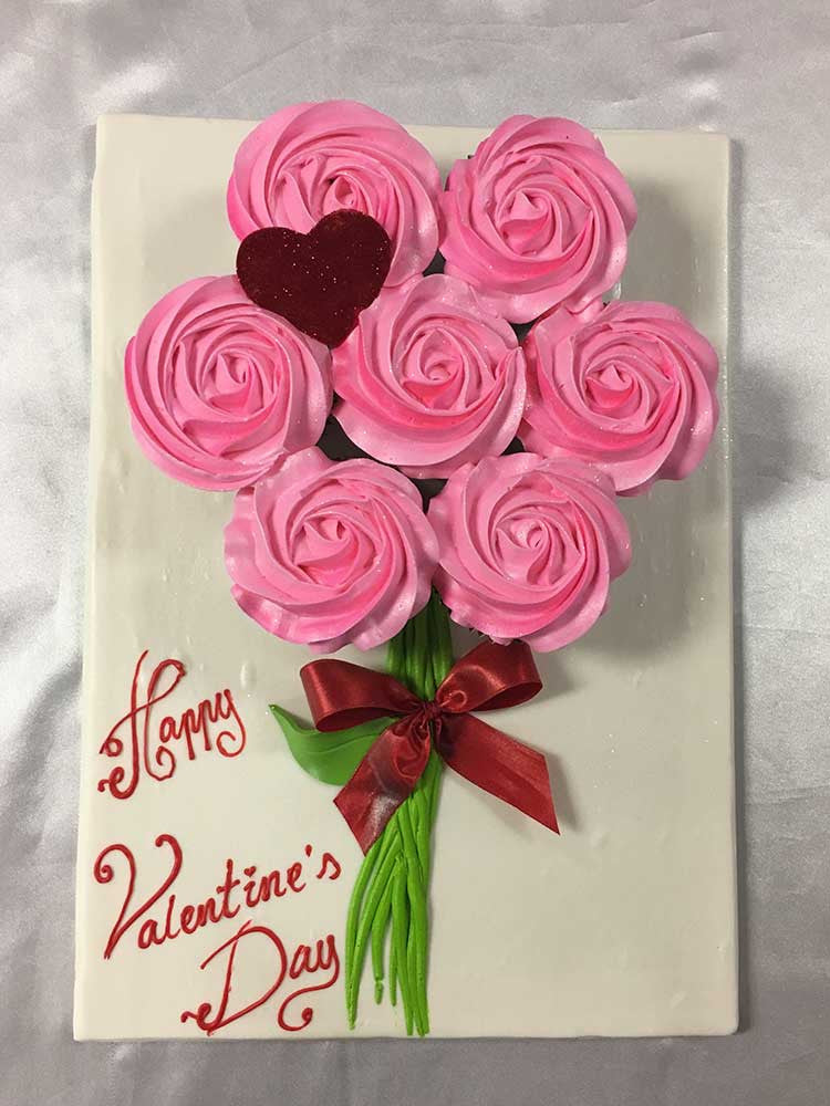 Seven pink buttercream cupcakes on a white board decorated beautifully like a floral bouquet. There is a red hear on top of one of the cupcakes. A "Happy Valentine’s Day" message is written on the board.