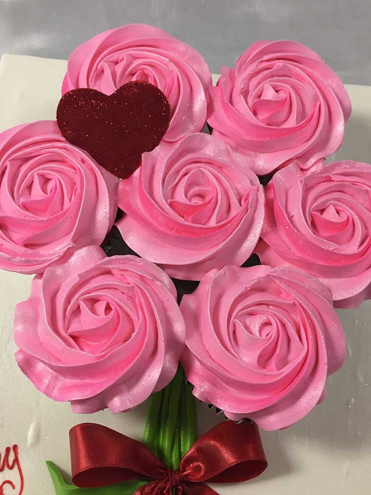 Seven pink buttercream cupcakes on a white board decorated beautifully like a floral bouquet. There is a red hear on top of one of the cupcakes.