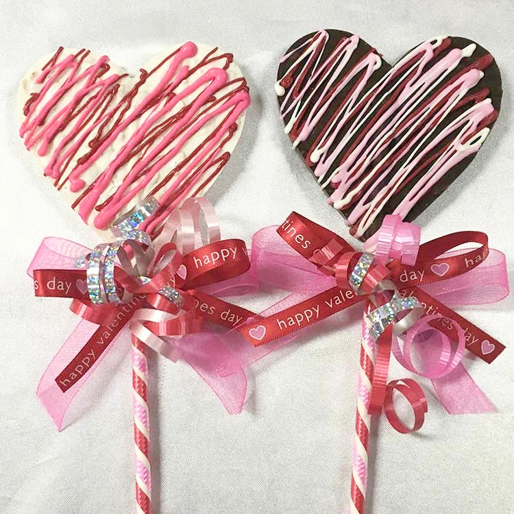 A white and brown crispy hearts next to each other, each on a 12” red, white, and pink striped lolly stick and tied with a red and pink bows.