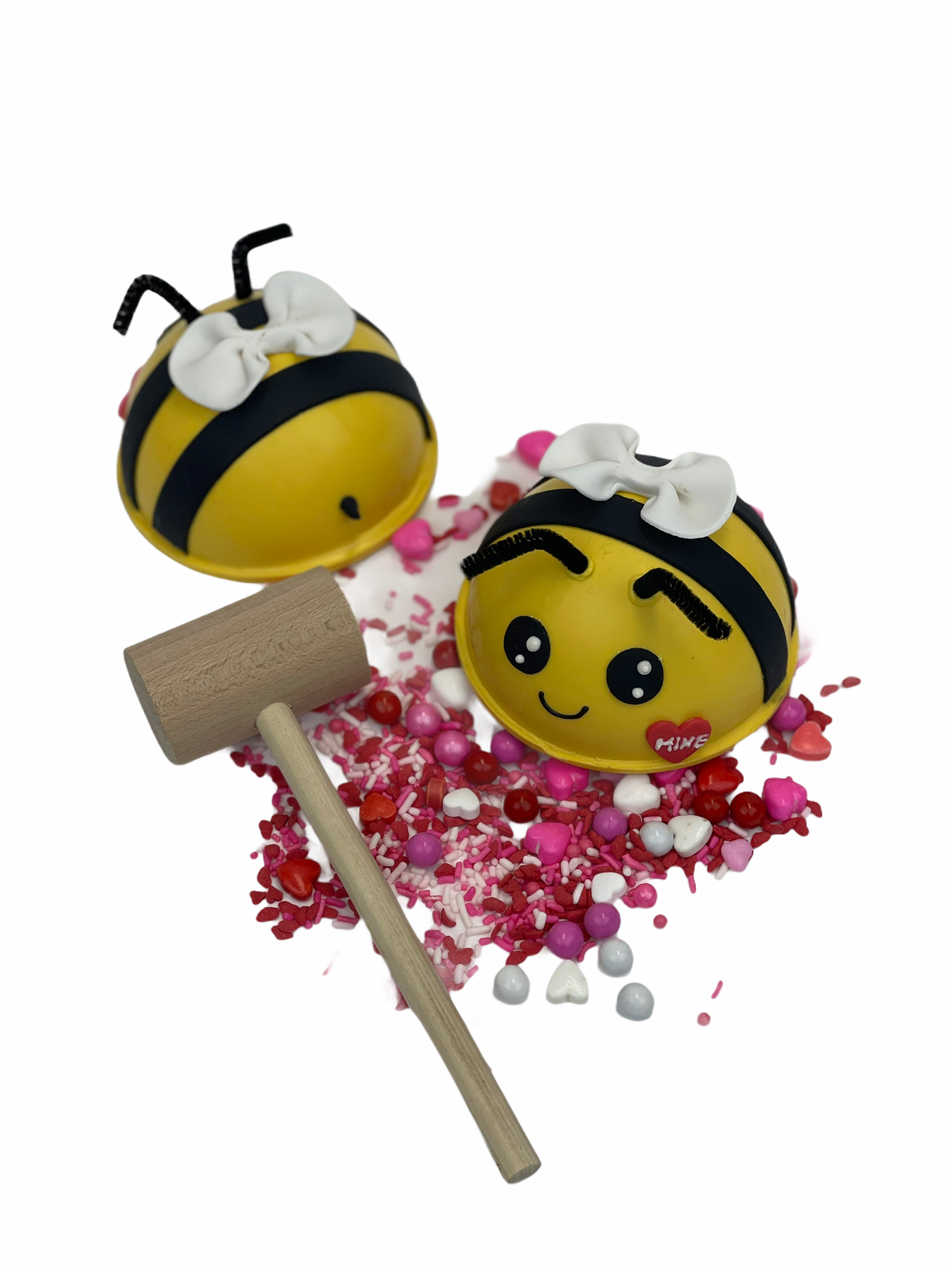 The back side of an unwrapped ” bee-shaped candy-filled chocolate sphere piñata next to the another unwrapped bee-shaped piñata (showing its front side) with a small heart on its face that says "Mine." There is a wooden mallet next to it.