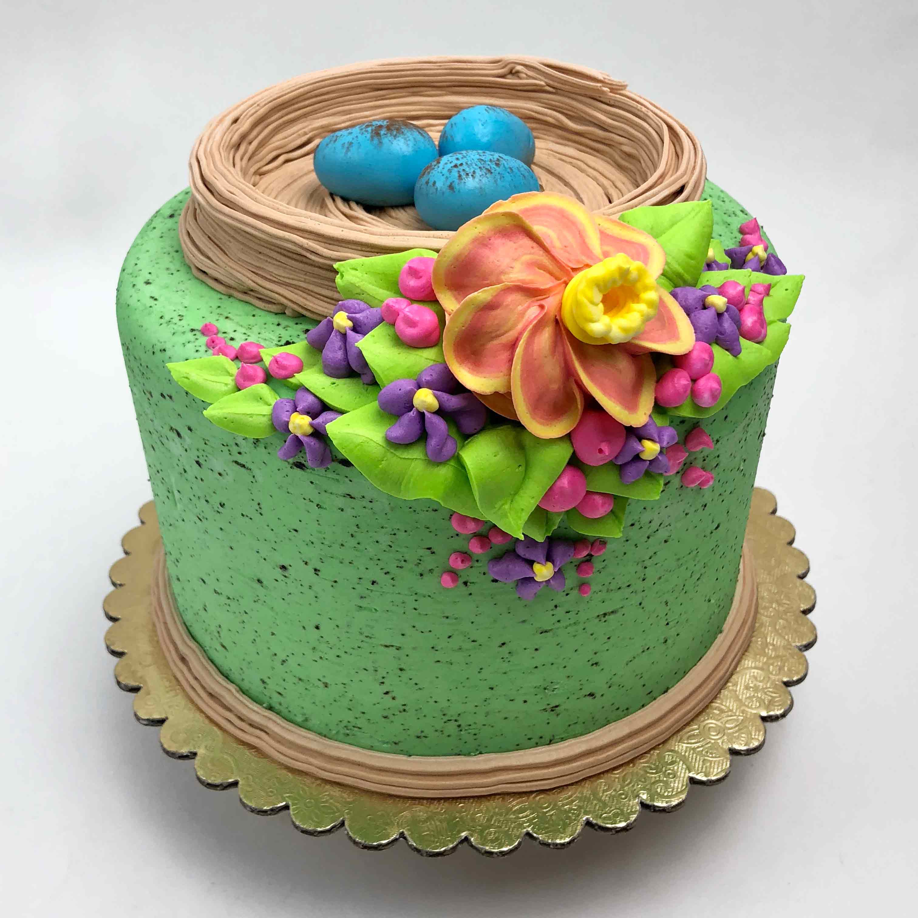 A green Easter Cake with a bird’s nest on top and three mini blue eggs inside the nest. The front of the cake is decorated with colorful flowers. The cake sits on a gold base.