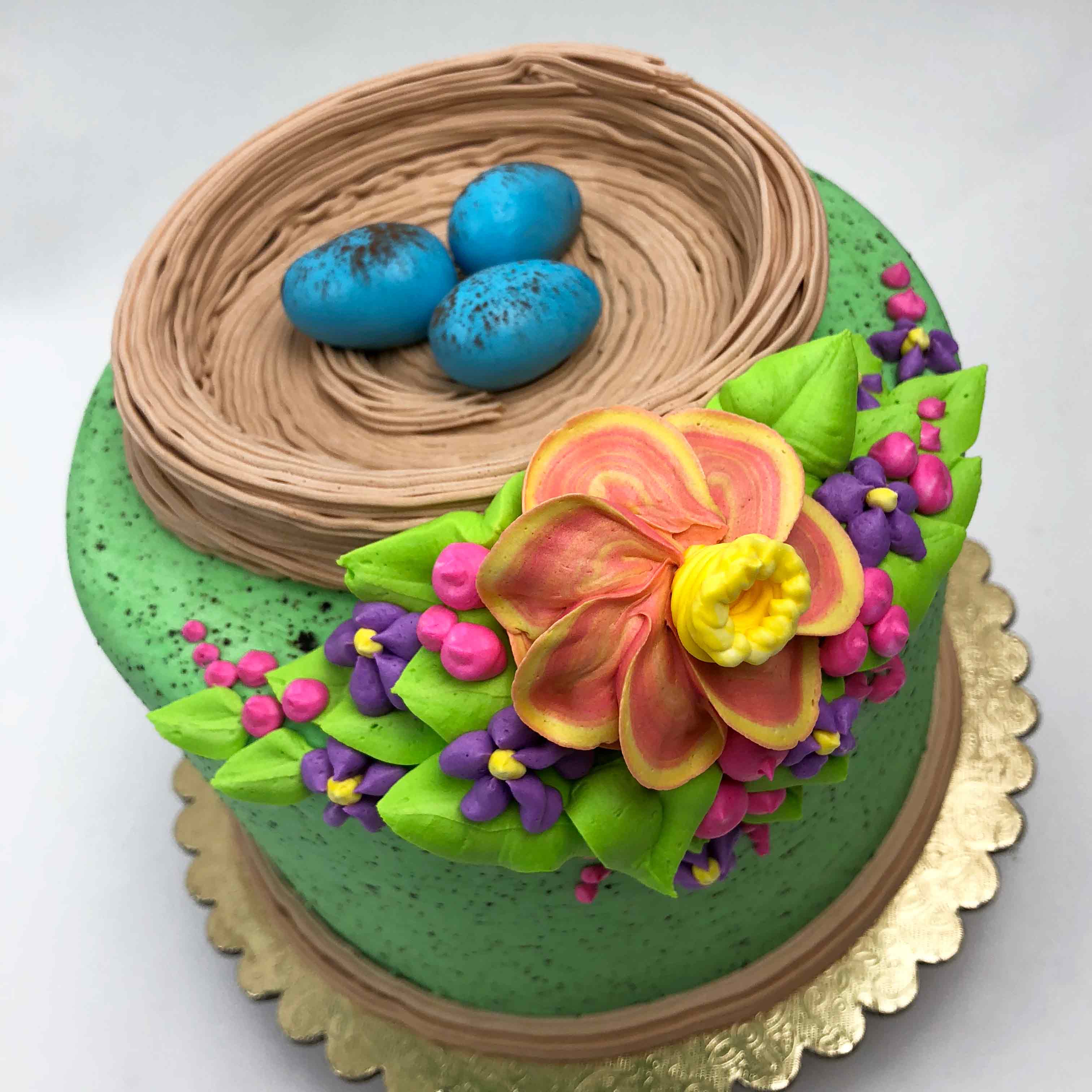 The top view of a green Easter Cake with a bird’s nest on top and three mini blue eggs inside the nest. The front of the cake is decorated with colorful flowers. The cake sits on a gold base.