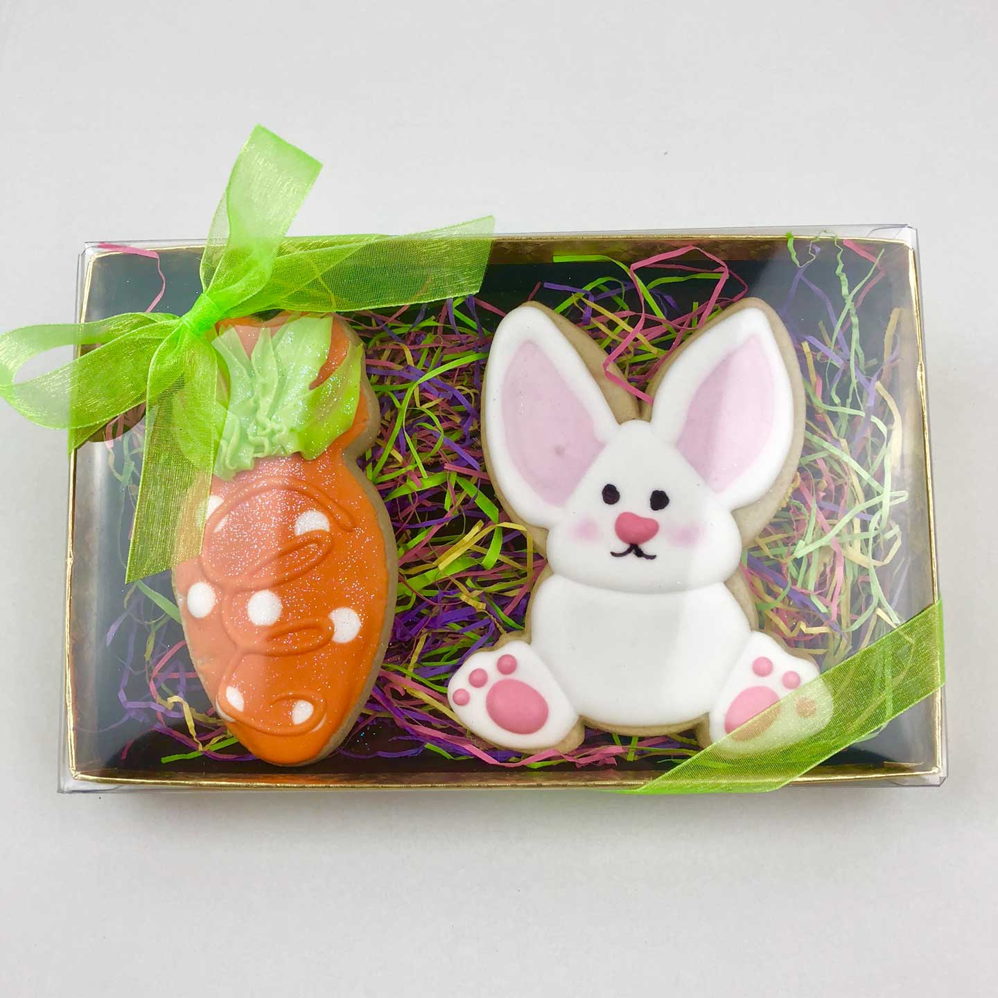 One orange carrot with white polka dots and a white bunny cookies in a box with colorful packing paper with a clear box top