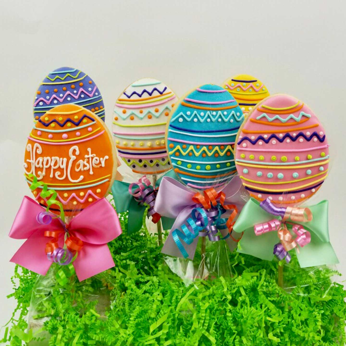 Six pink, blue, yellow, white, orange, and purple colors decorated Easter egg sugar cookies on sticks. The orange cookie features a white color "Happy Easter" message and a pink bow. The cookies are displayed on top of green crinkled packing paper.