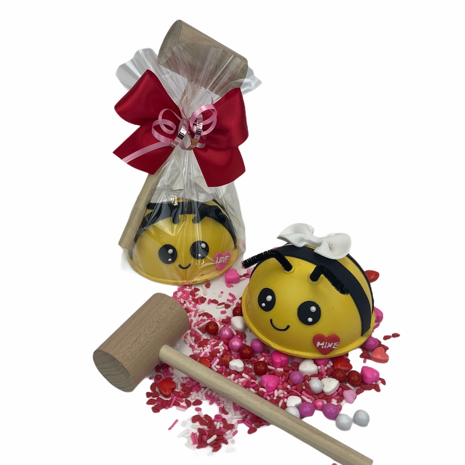 A 4” bee-shaped, candy-filled chocolate sphere piñata that has a small red heart on its face that says "love," wrapped in clear cellophane, has a mallet and is tied with red and pink bows. The other piñata is not wrapped. It has a small red heart on its face that says "Mine." There is a wooden mallet next to it.