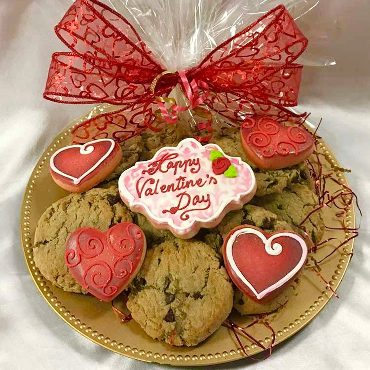 A few gourmet chocolate chip cookies, one large decorated sugar cookie that has "Happy Valentine's Day" message on it, and four smaller decorated red heart-shaped sugar cookies on gold platter, wrapped in clear cellophane and tied with a big red bow.