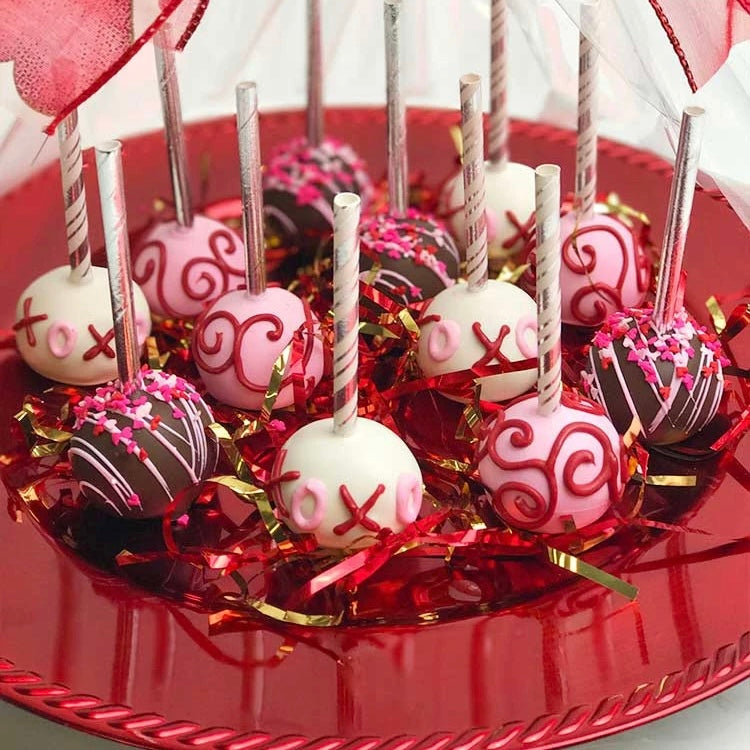 A platter of 12 Valentine's Cake Pop in an assortment of brown, white, and pink designs, wrapped with clear cellophane and a Valentine's bow with red hearts.