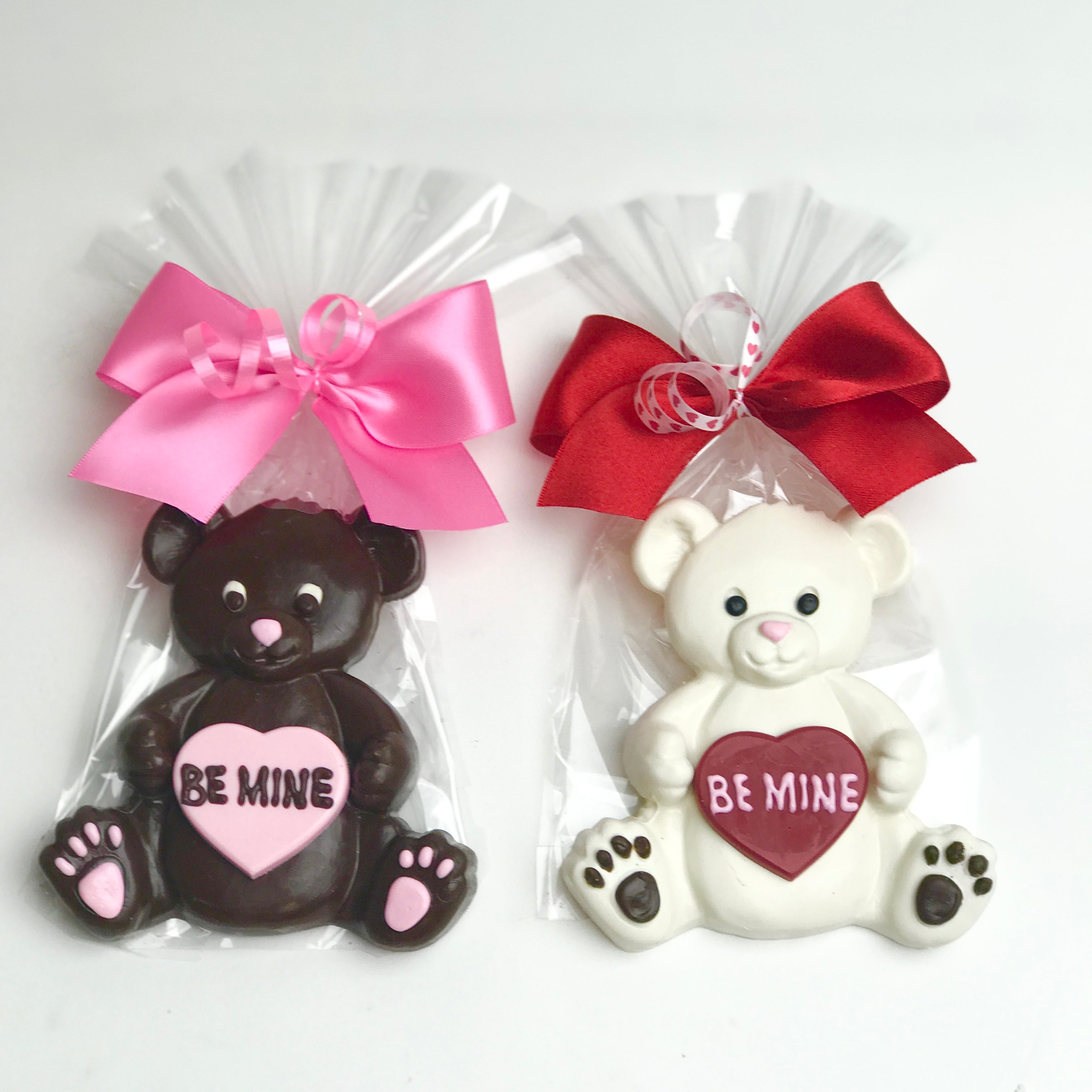 A brown chocolate bears holding a pink heart with the message "Be mine" written on it. The bear is wrapped in clear cellophane and tied with pink satin bows. Next to it there is a a white chocolate bears holding a red heart with the message "Be mine" written on it. The bear is wrapped in clear cellophane and tied with red and white satin bows.