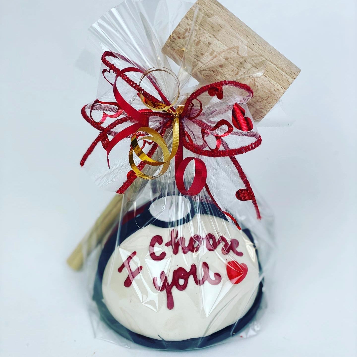 A white, red, and black 4” candy-filled chocolate piñata with the saying ""I Choose You" written on it. It is wrapped in clear cellophane with a wooden mallet and red and gold ribbons.