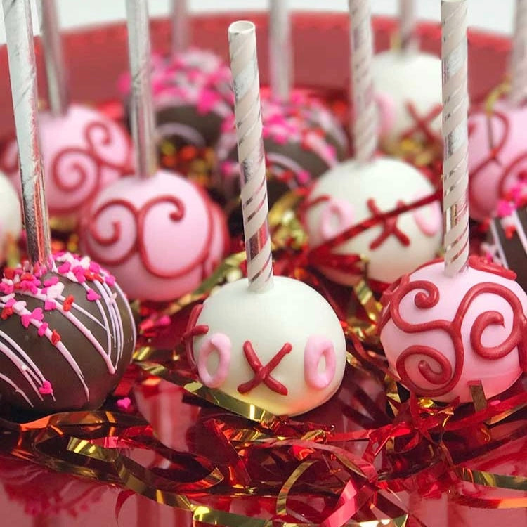 A platter of 12 Valentine's Cake Pop in an assortment of brown, white, and pink designs, wrapped with clear cellophane and a Valentine's bow with red hearts.