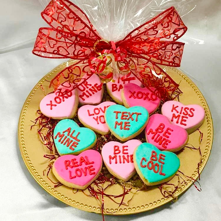 A dozen of ink and teal color heart-shaped sugar cookies decorated with fun Valentine’s Day sayings like "XOXO", "Be Mine", "Be Cool", "All Mine" "Real Love", "Kiss Me", "Love You", "UR Mine" and "Text Me" on them and wrapped on a platter in clear cellophane and tied with a beautiful red bow.