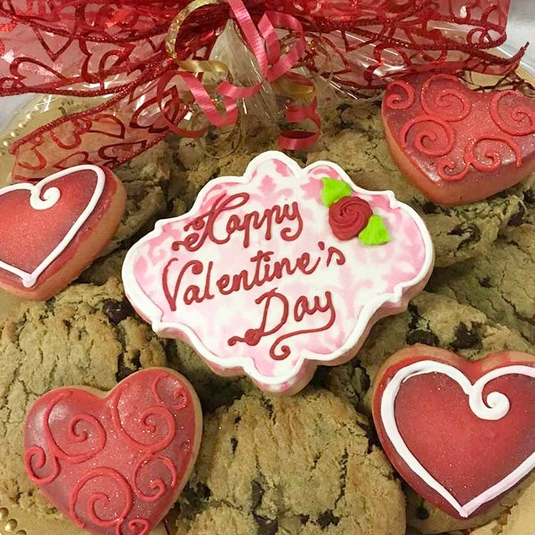 A few gourmet chocolate chip cookies, one large decorated sugar cookie that has "Happy Valentine's Day" message on it, and four smaller decorated red heart-shaped sugar cookies on gold platter, wrapped in clear cellophane and tied with a big red bow.