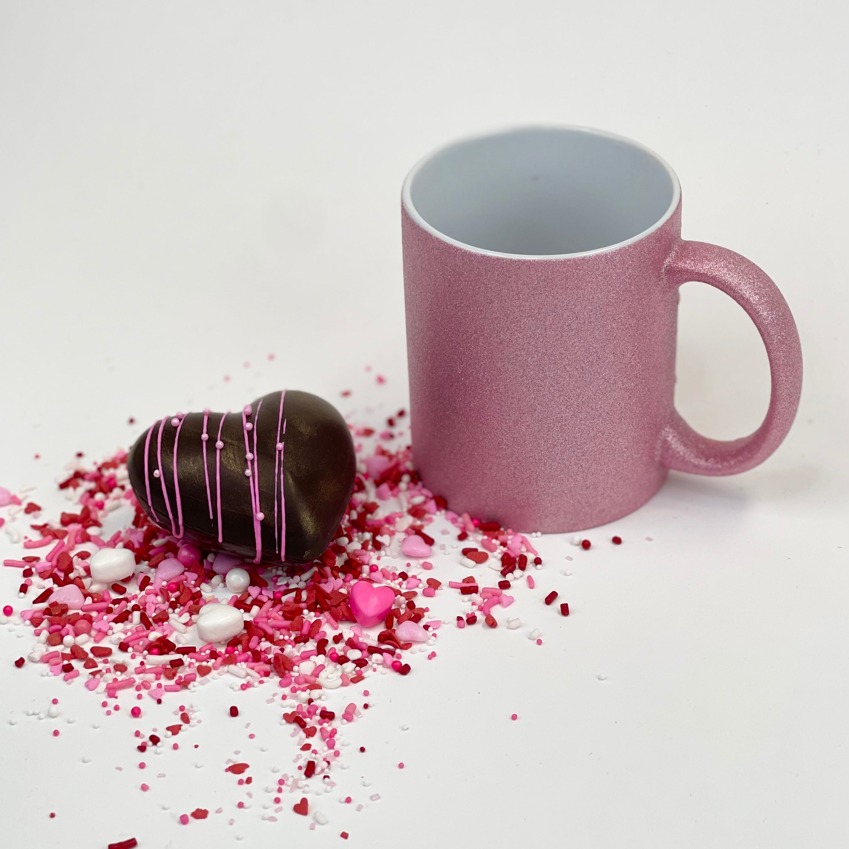 A Valentine’s heart hot cocoa bomb with pink drizzles next to a pink mug