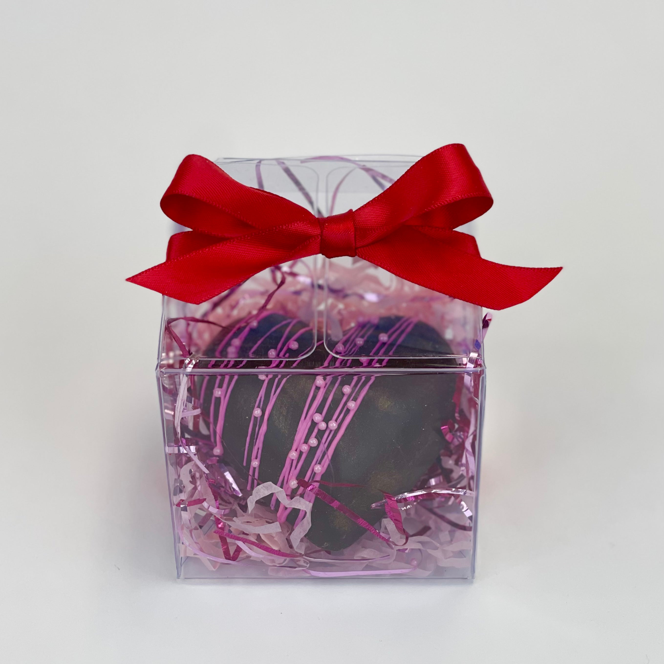 A Valentine’s heart hot cocoa bomb with pink drizzles is inside a clear box with crinkle paper. There is a red bow on top of the box.